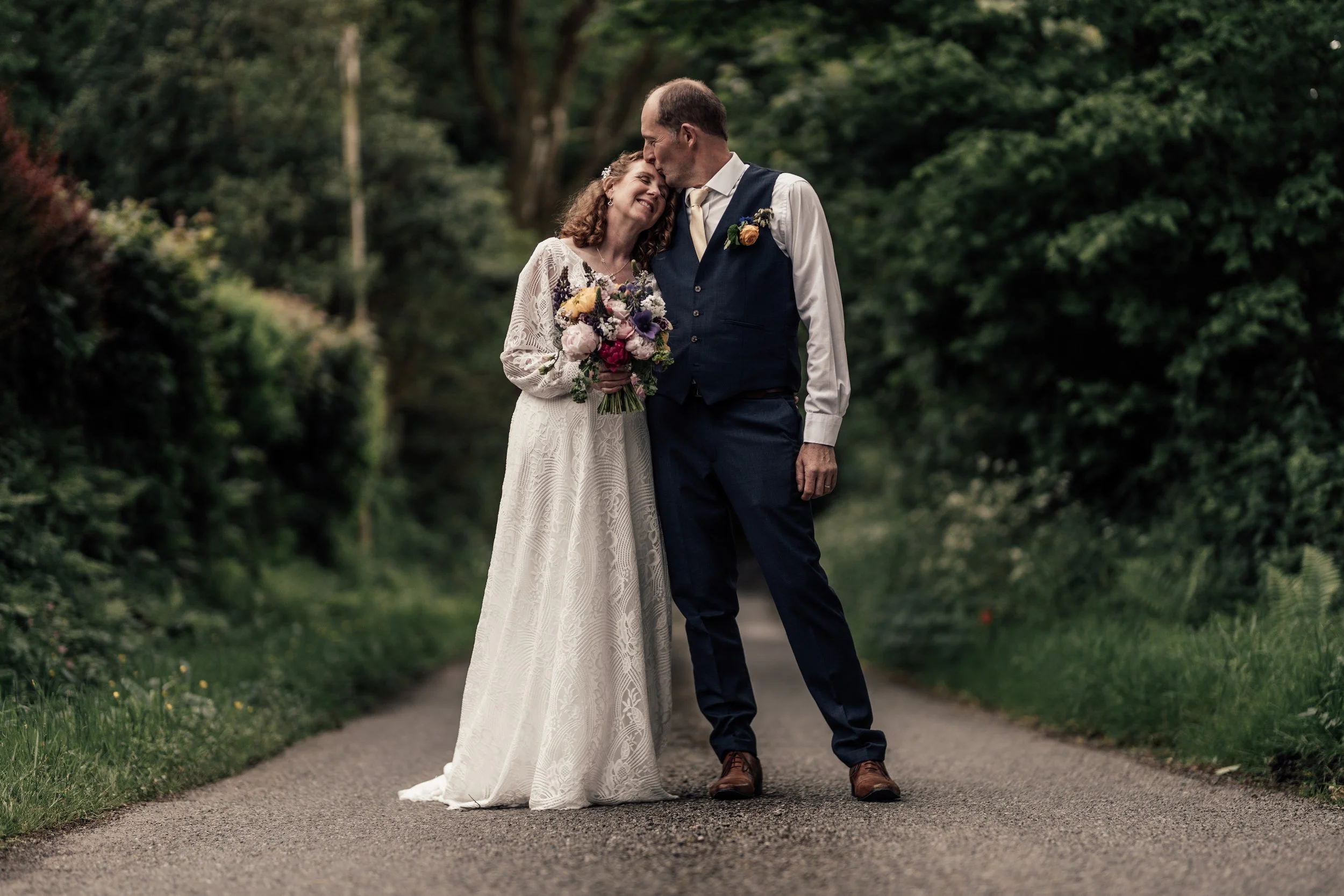 A bride and groom standing on a quiet pathway surrounded by lush green trees, sharing a tender moment with the bride holding a colorful bouquet of flowers and the groom wearing a dark suit with a boutonniere.