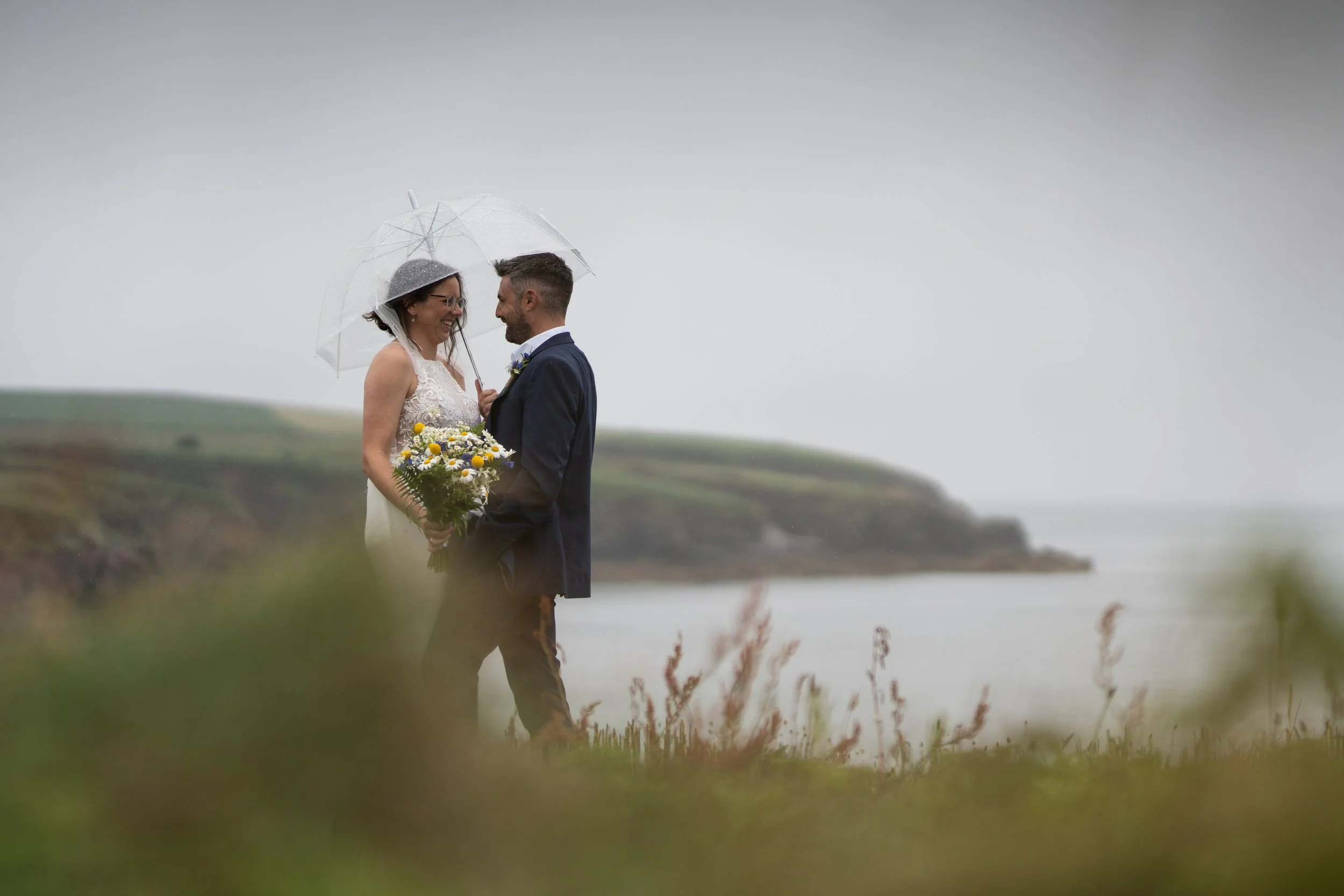 A couple in wedding attire stands close under a transparent umbrella on a rainy day near a body of water, with a grassy landscape and cliffs in the background. The bride holds a bouquet of yellow and white flowers.