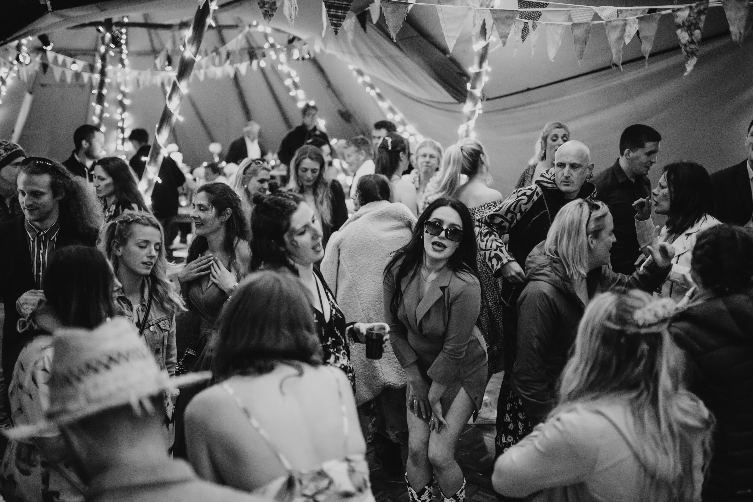 People dancing and socializing inside a decorated tent at a party or celebration, illuminated with string lights and bunting.