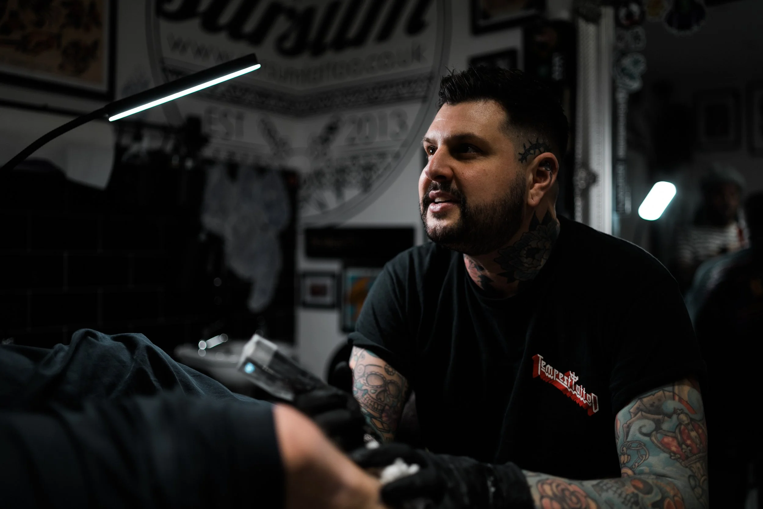 A man with tattoos on his arms and neck, short dark hair, and facial hair, sits in a tattoo studio. He is wearing a black T-shirt with red and white writing. The studio has dark walls, framed pictures, and a large circular artwork on the wall. A tatt