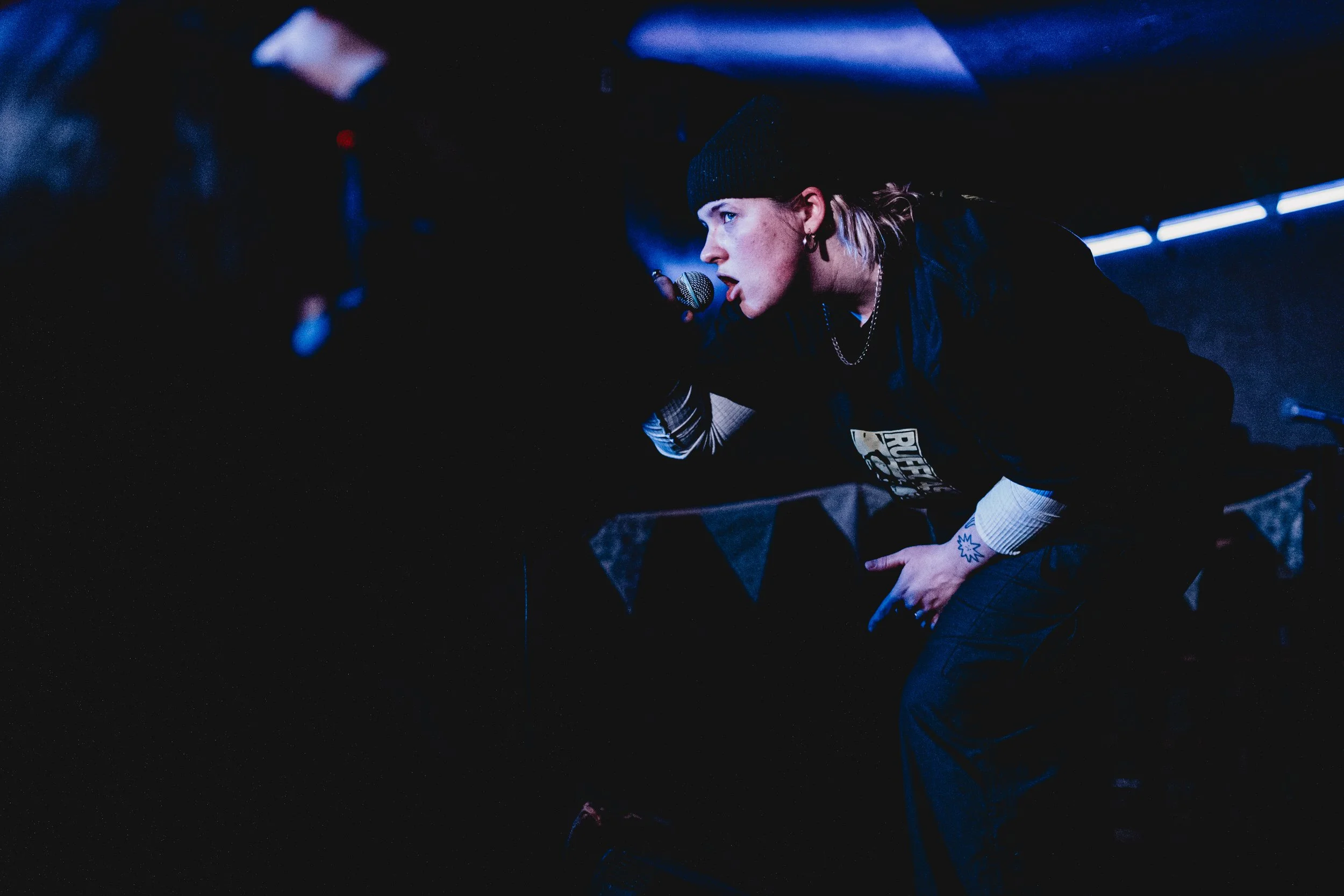 A woman wearing a black beanie and dark clothing is performing on stage, holding a microphone close to her face, with stage lights illuminating her profile. Live event photography.