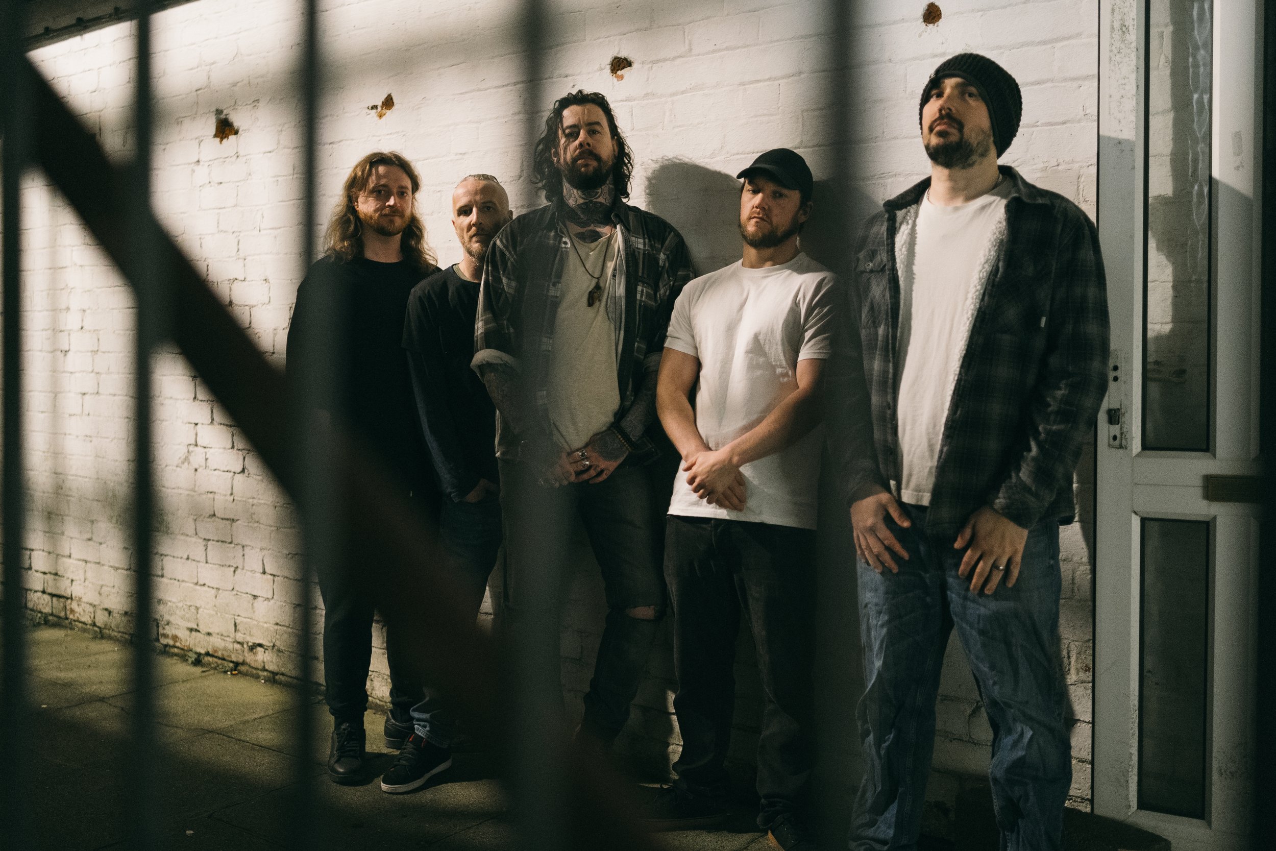 Five men standing against a white brick wall, in a dimly lit urban setting. Promotional Music Photography.