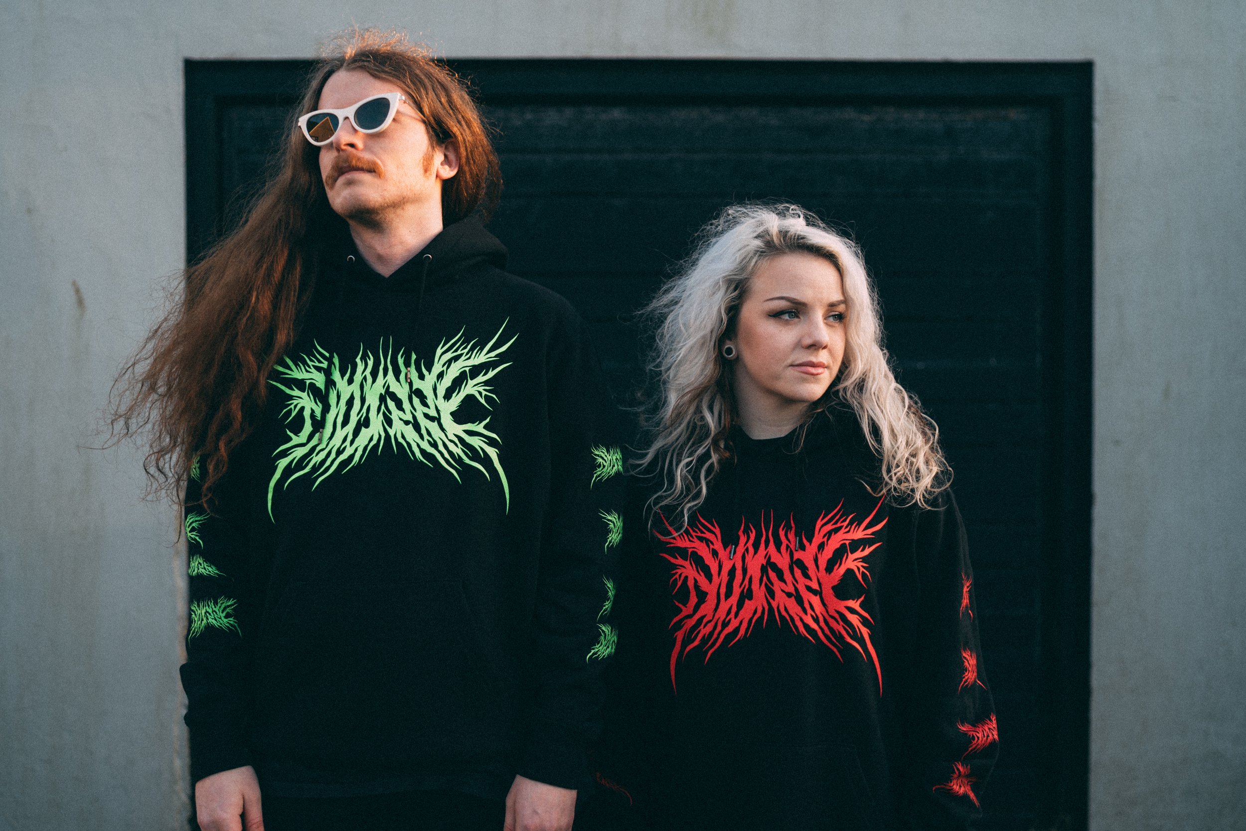 Two young adults standing in front of a black garage door, wearing black hoodies with green and red graphic designs and sunglasses. NOIZZE Logo Merch.