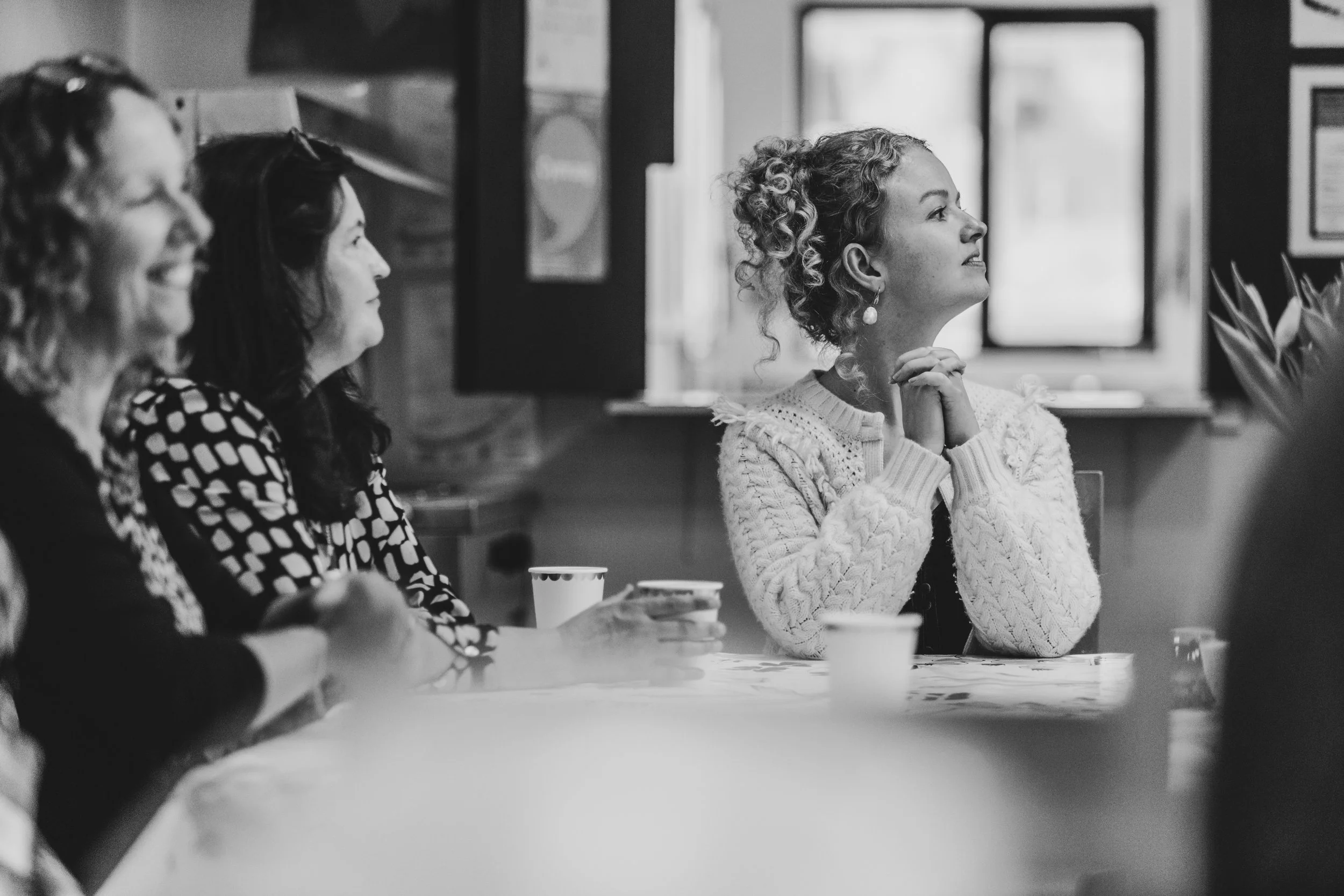 A woman with curly hair and earrings sitting at a table with her hands clasped, attentively listening, with two women partially visible beside her, in a room with windows and framed pictures on the wall.