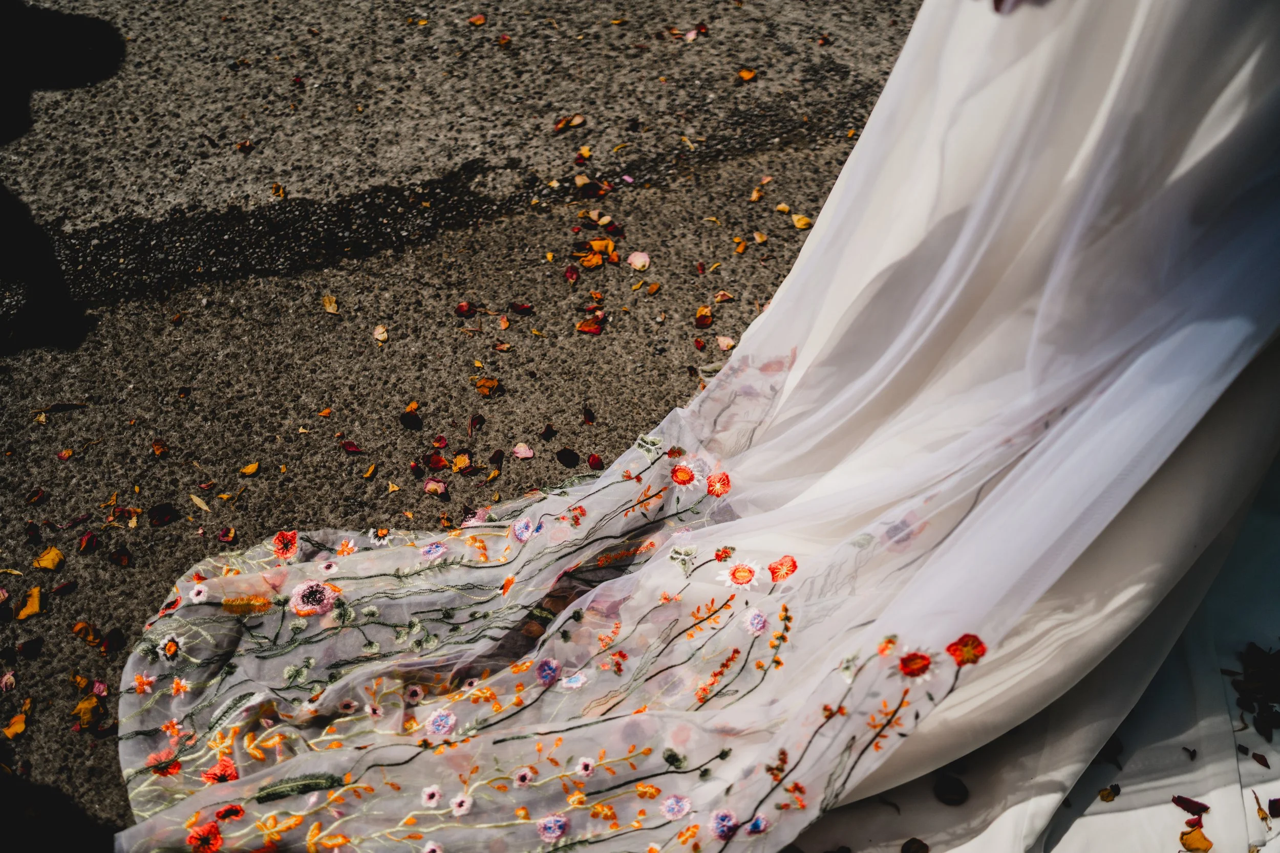 A white dress with colorful floral embroidery lying on the ground with flower petals scattered around.