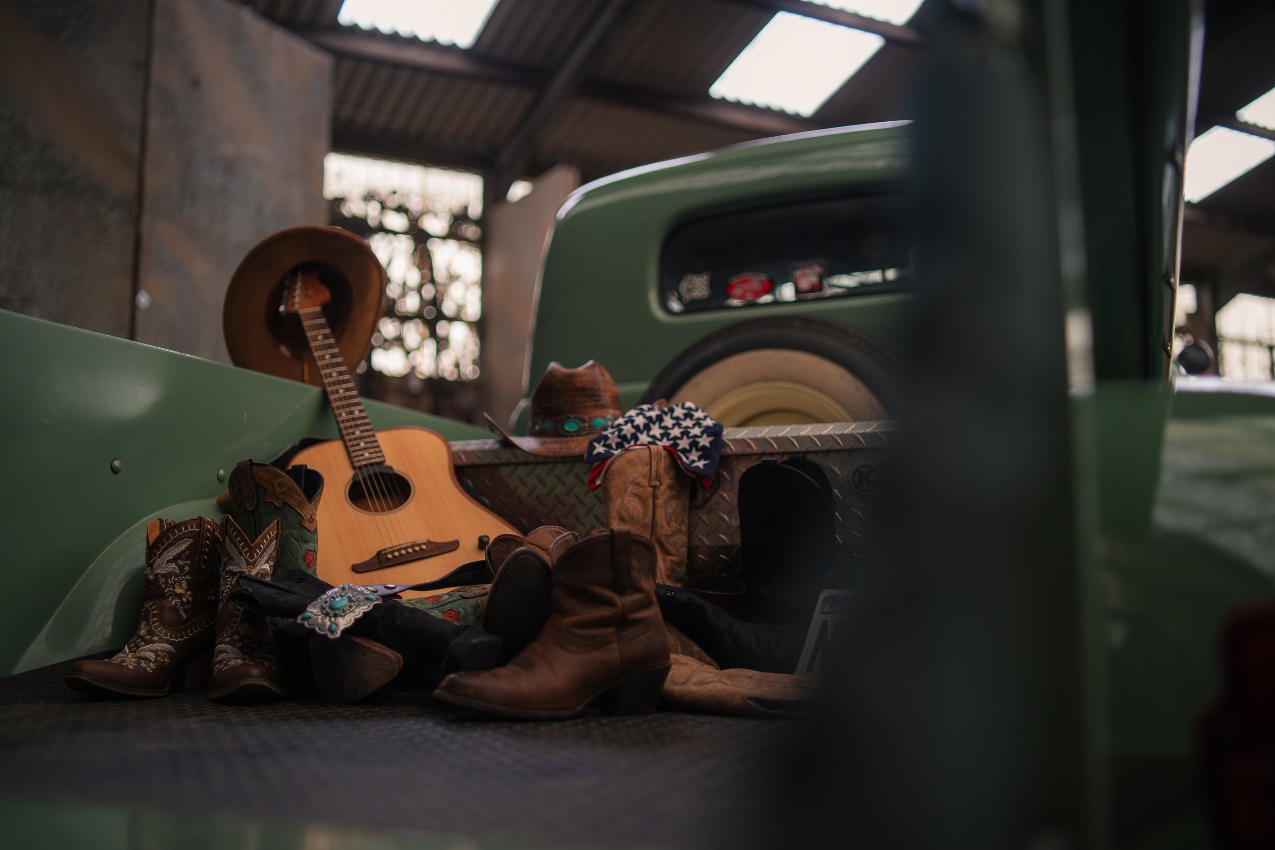 A collection of cowboy boots, a guitar, a cowboy hat, a bandana, and sunglasses in the bed of a green pickup truck inside a barn.
