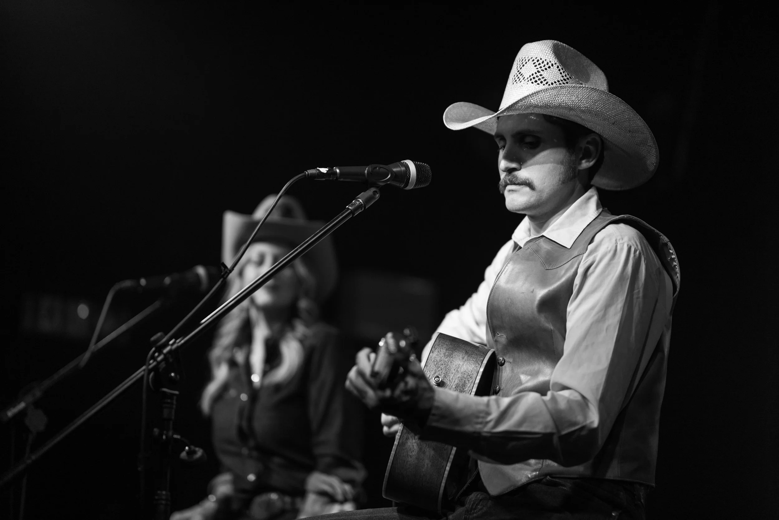 A man wearing a cowboy hat and Western-style clothing playing an acoustic guitar and singing into a microphone on stage, with a woman in the background also wearing a cowboy hat. Jesse Daniel Country Artist.