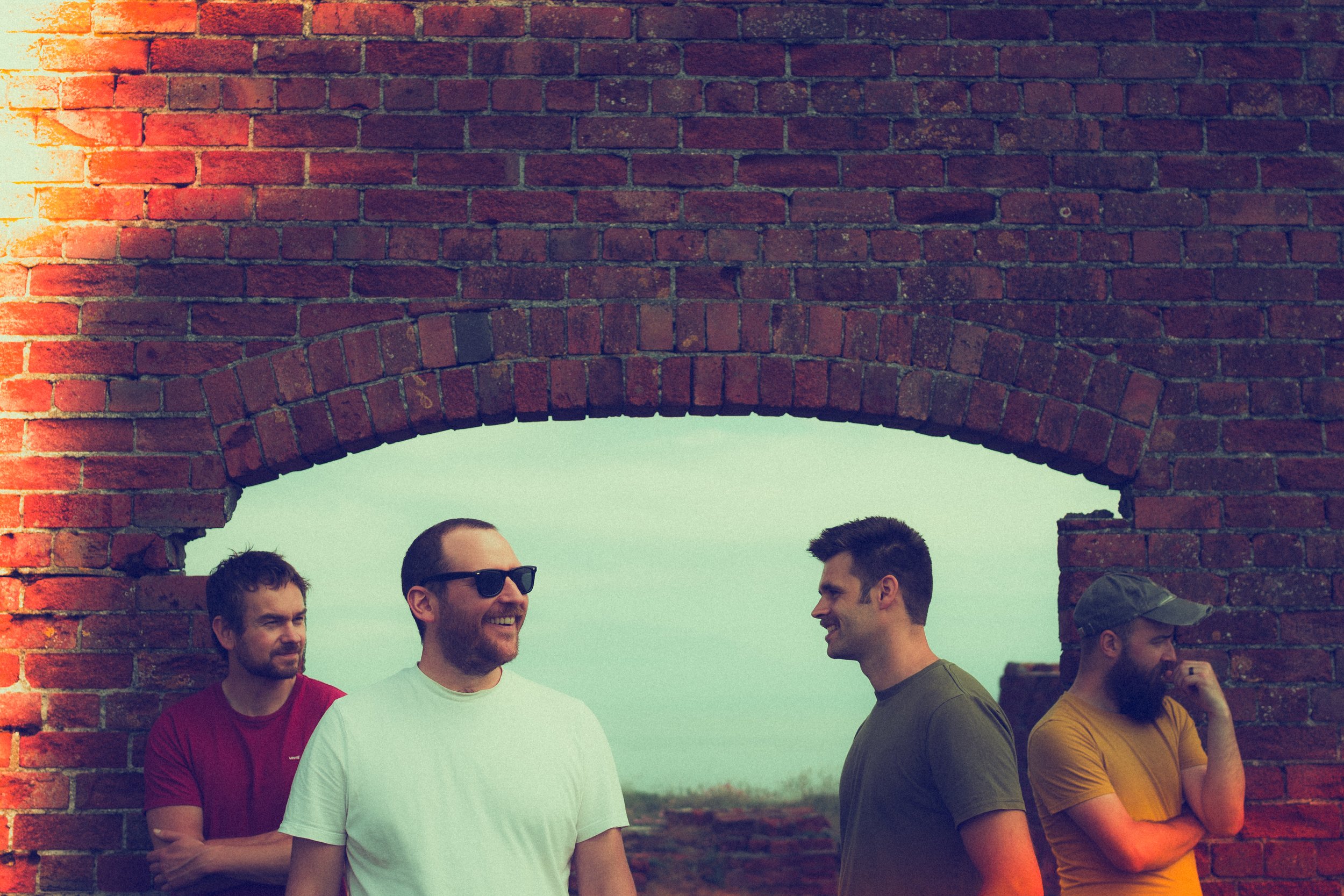 Four men standing outdoors near a brick wall with a large archway, smiling and talking, during daytime. Promotional Music Photography.
