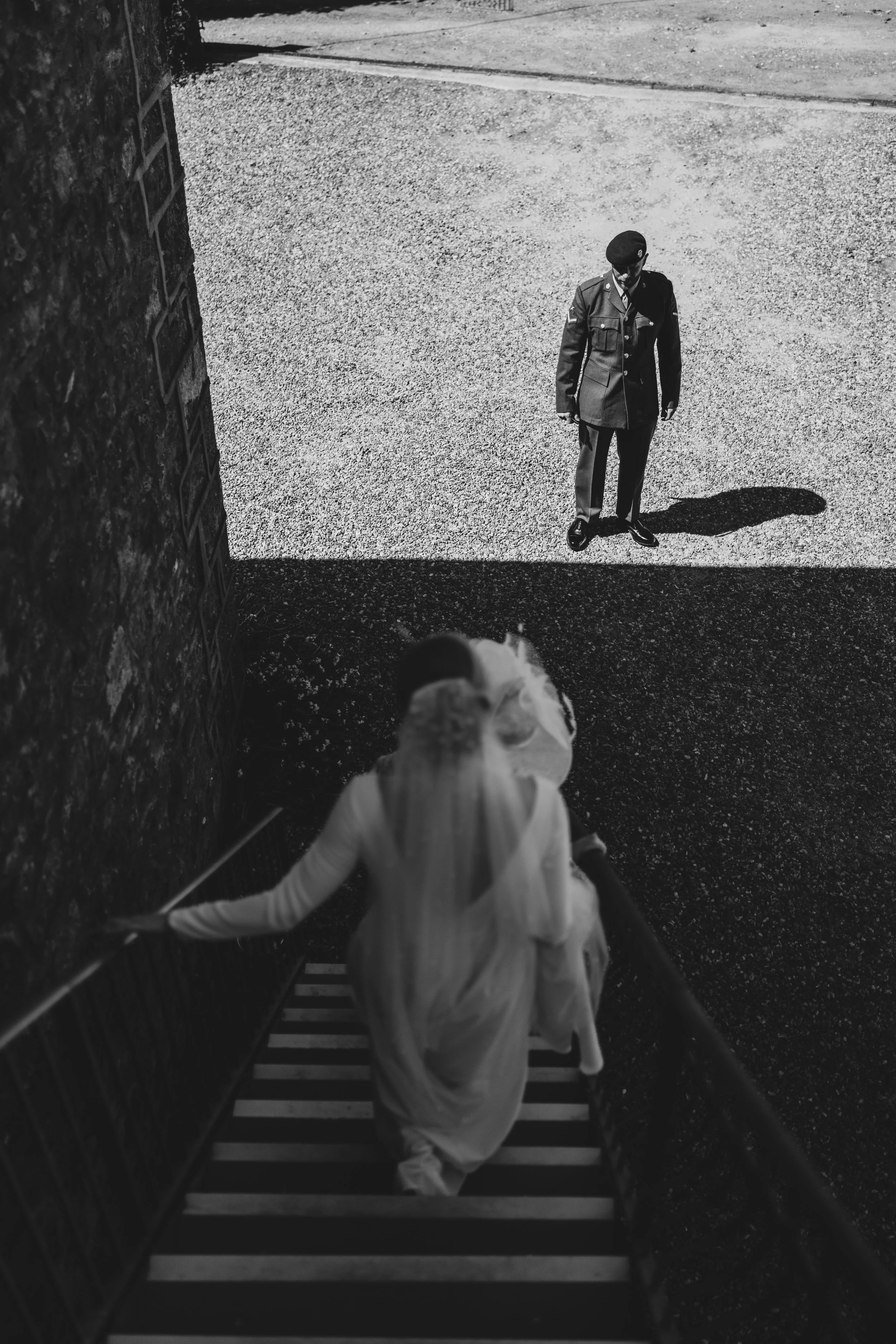 A bride in a wedding dress and veil walking up outdoor stairs, with a man in a military uniform standing at the bottom, outside of a brick building, in black and white.