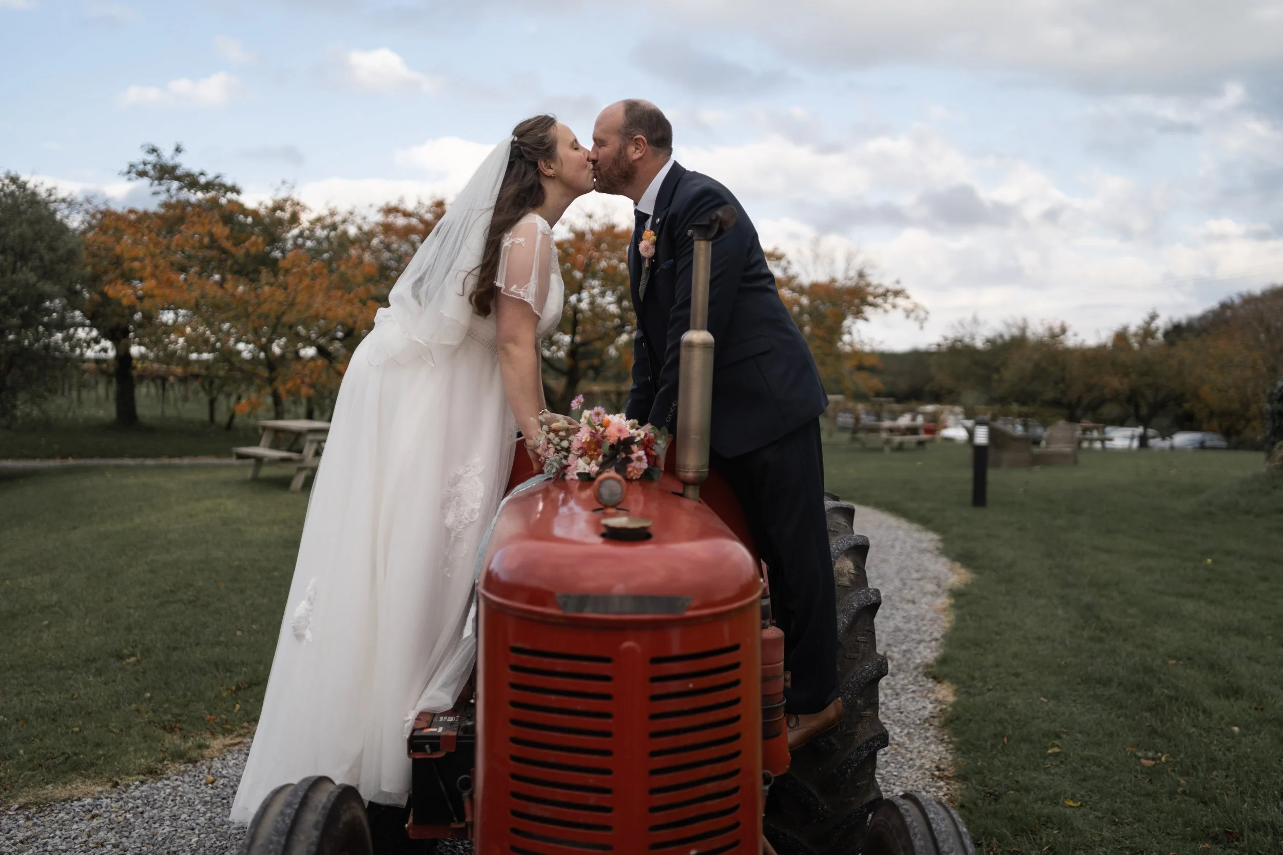 Bride and groom kissing on a vintage tractor during a wedding photoshoot in an outdoor park with trees showing fall colors.