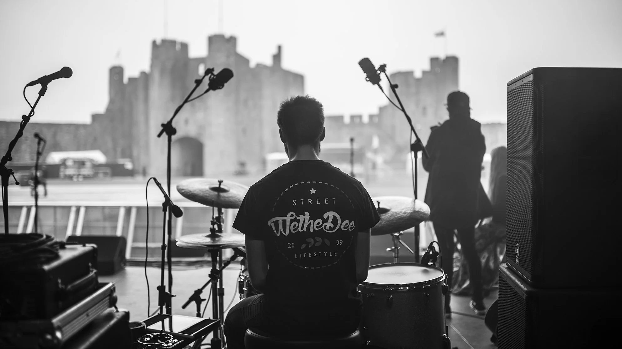 A person sitting behind a drum set on a stage, facing a castle-like structure outside. Other musicians are in the background, and microphones and speakers surround the stage.