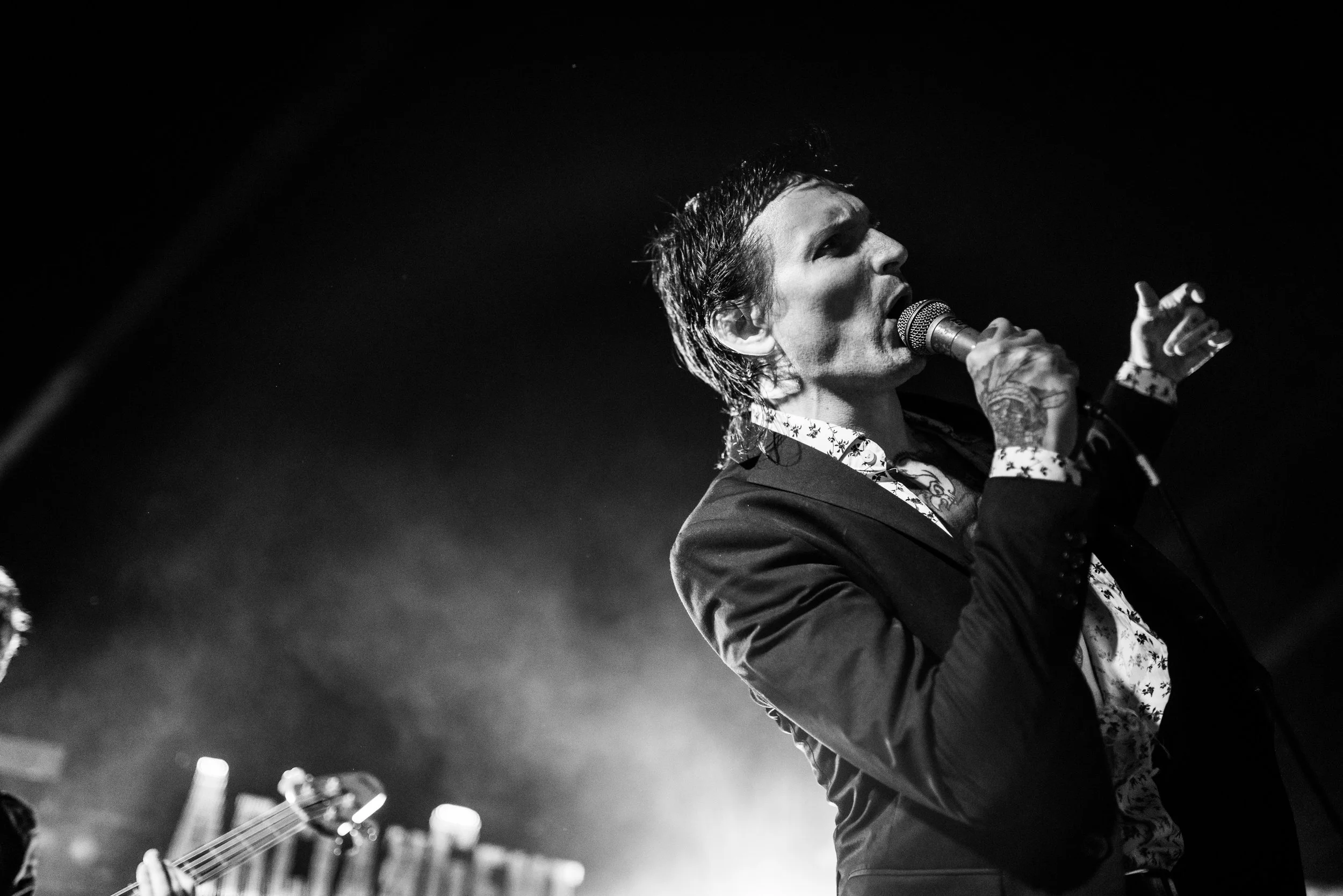 A male singer with wet hair holding a microphone and singing on stage, with a guitarist partially visible in the background, in black and white. Festival Photography.