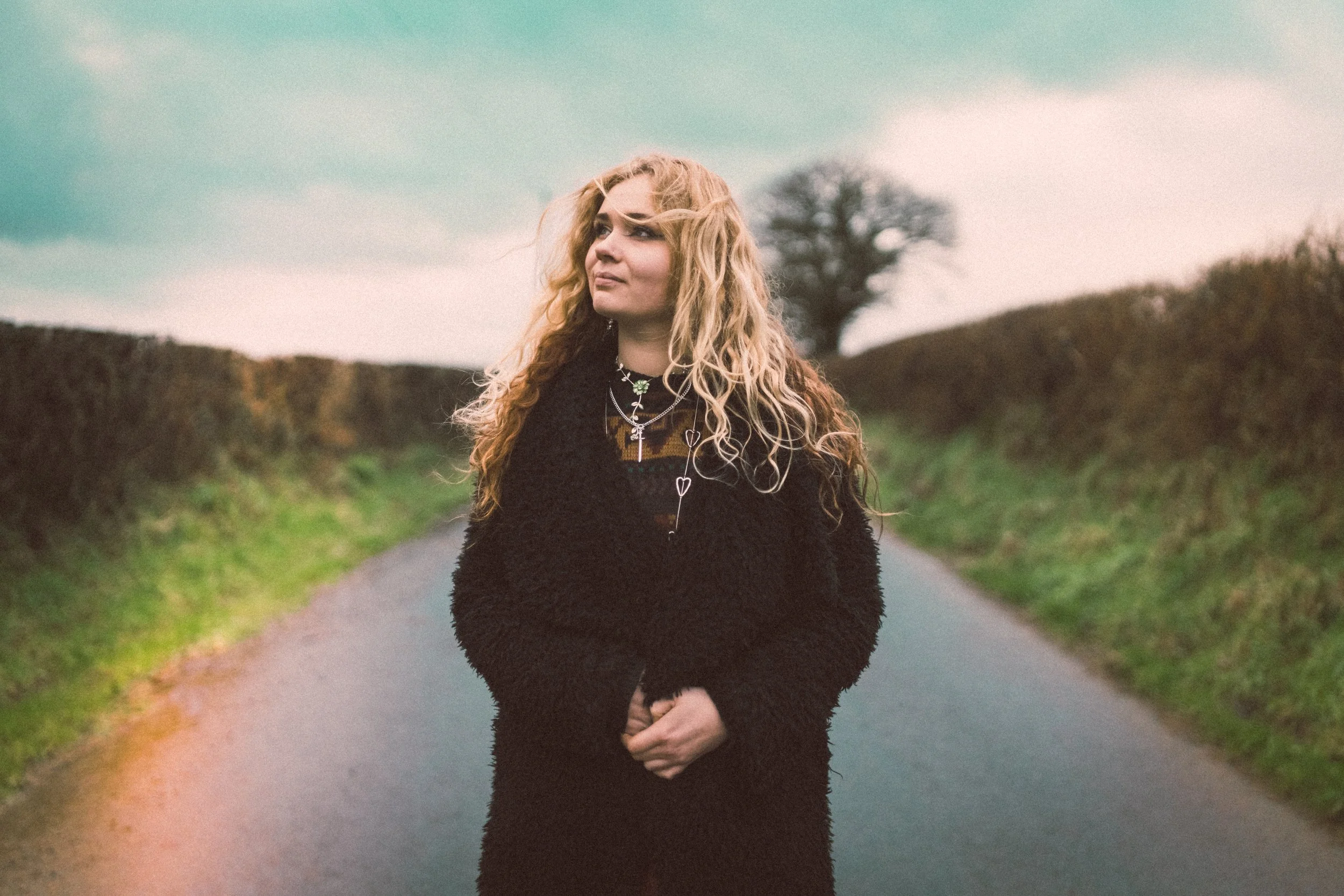 A young woman with curly blonde hair standing on a rural road with hedges on both sides and a tree in the background under a cloudy sky.