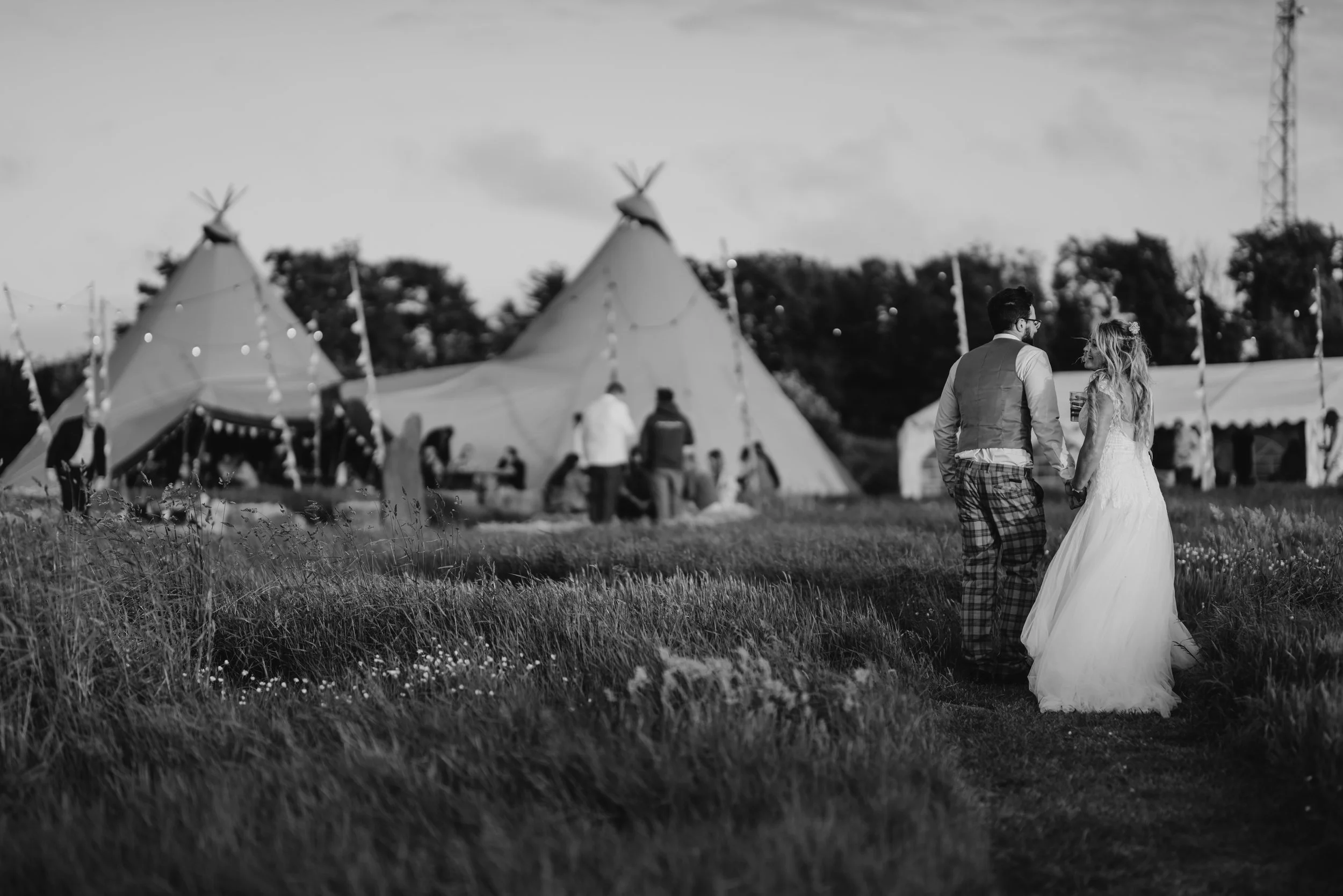 A black and white photo of a bride and groom walking hand in hand in a field with tents in the background, possibly at a wedding or outdoor event.