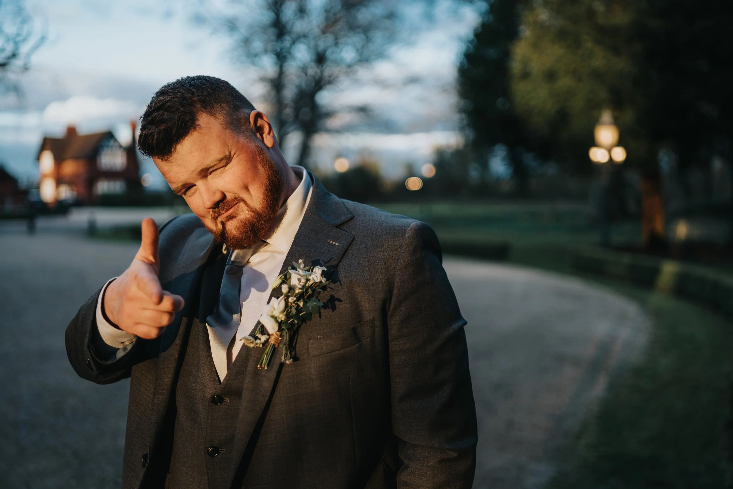 A man in a gray suit with a boutonniere making a finger gun gesture and winking outdoors during evening.
