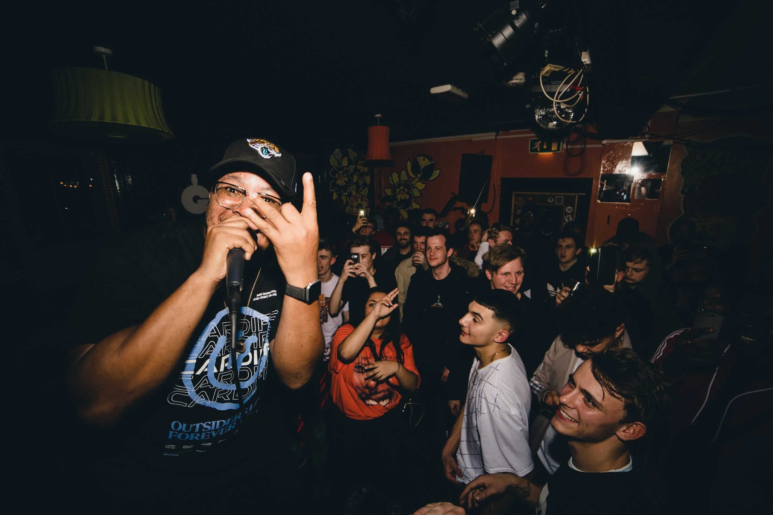 A male performer is on stage holding a microphone and making a hand gesture, wearing a black cap and glasses, while an audience of young people is dancing and smiling in a lively indoor Grime event.