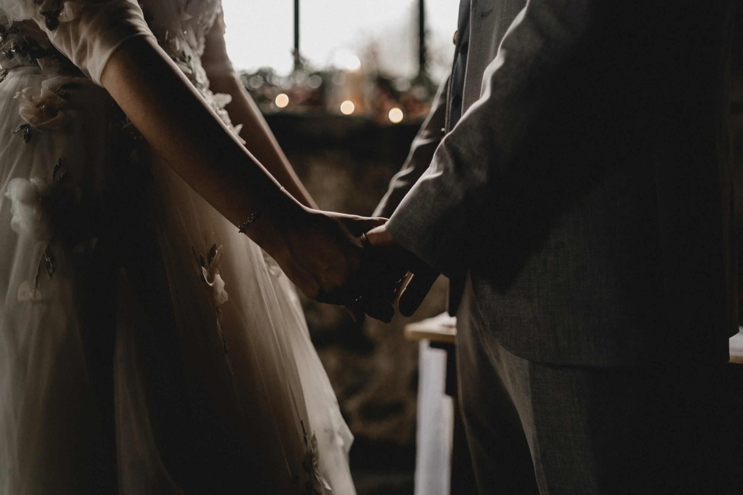A couple holding hands during a wedding ceremony, with soft lighting and the bride in a dress with floral embellishments. Alt Wedding. Alternative wedding photography.