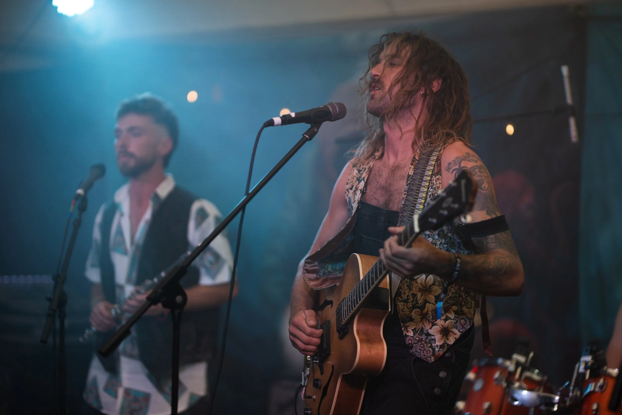 Musicians performing on stage, with one playing guitar and singing into a microphone and another playing a clarinet in the background, under dim stage lighting.