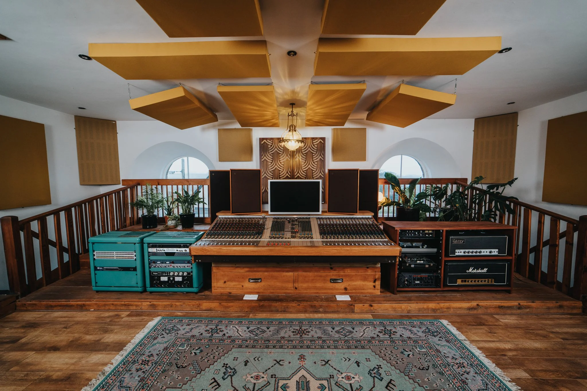 Recording studio with wooden flooring, mixing console, speakers, and musical equipment, plants, and decorative acoustic panels on the walls and ceiling.