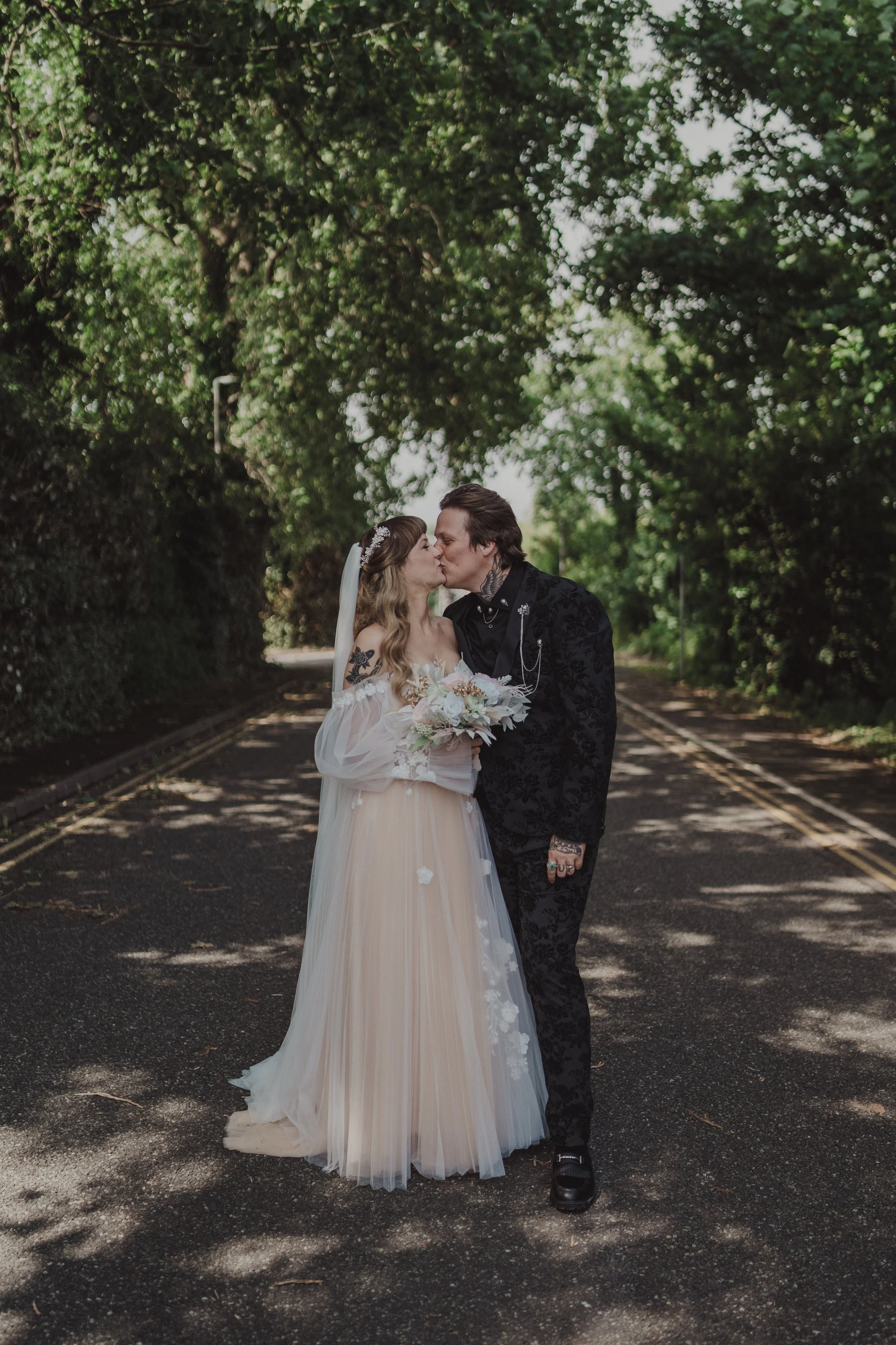 A bride and groom share a kiss on a tree-lined street during their wedding. The bride wears a white gown with flowing skirt, and the groom wears a black suit with gothic style accessories.