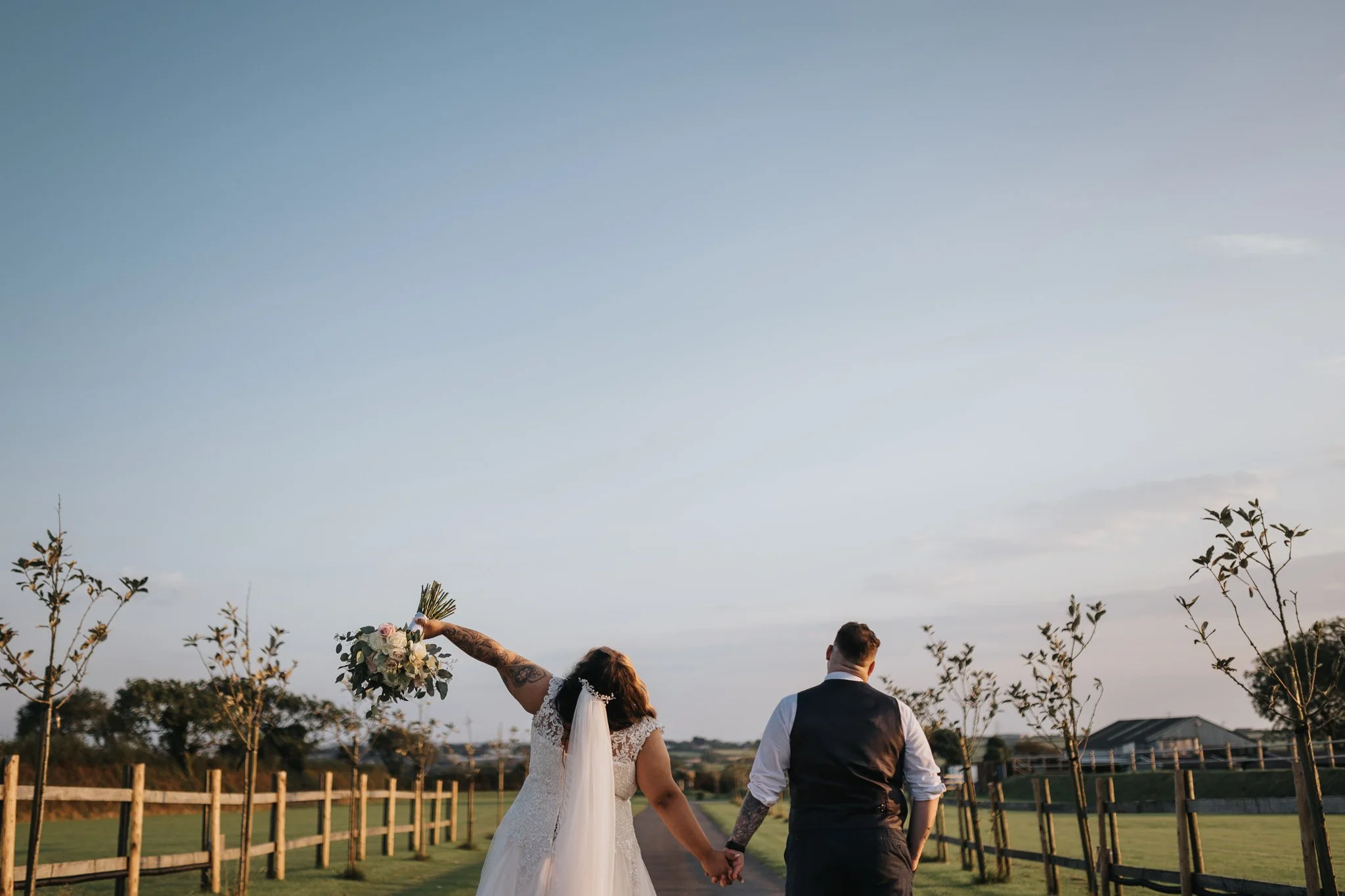 A bride and groom hold hands and walk down a path outdoors at sunset, the bride holding a bouquet and raising her other arm, with small trees and a fence along the path.