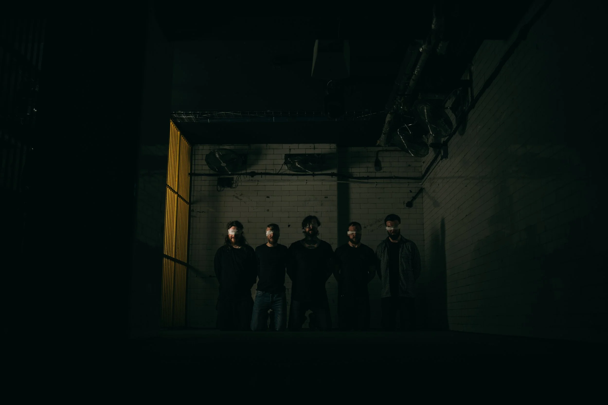 Five people stand in a dark, industrial space with brick walls, all wearing black and masks, illuminated by minimal lighting. Promotional music photography.