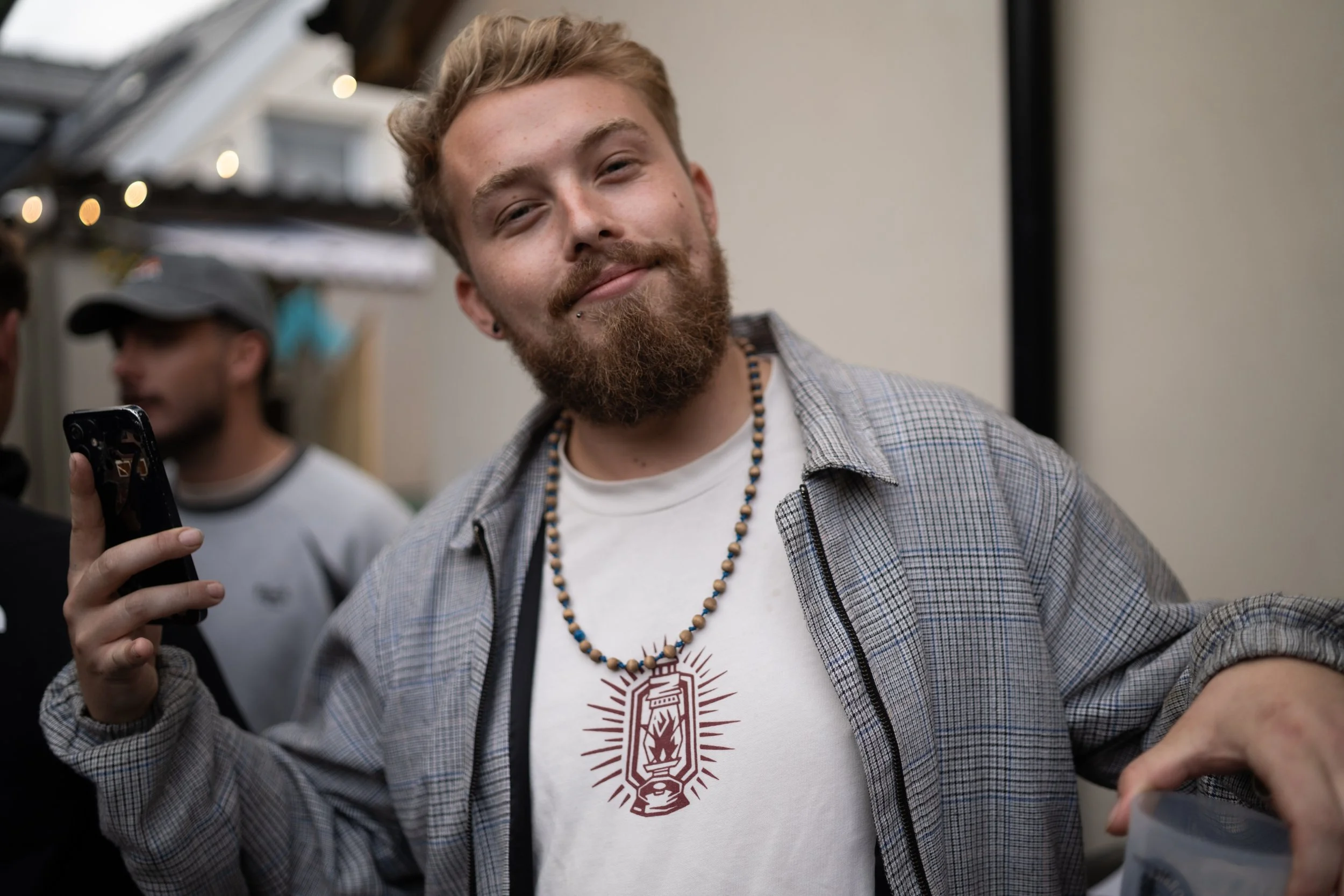 Young man with a beard and piercings smiling while holding a phone at a social gathering, wearing a necklace and a casual shirt.