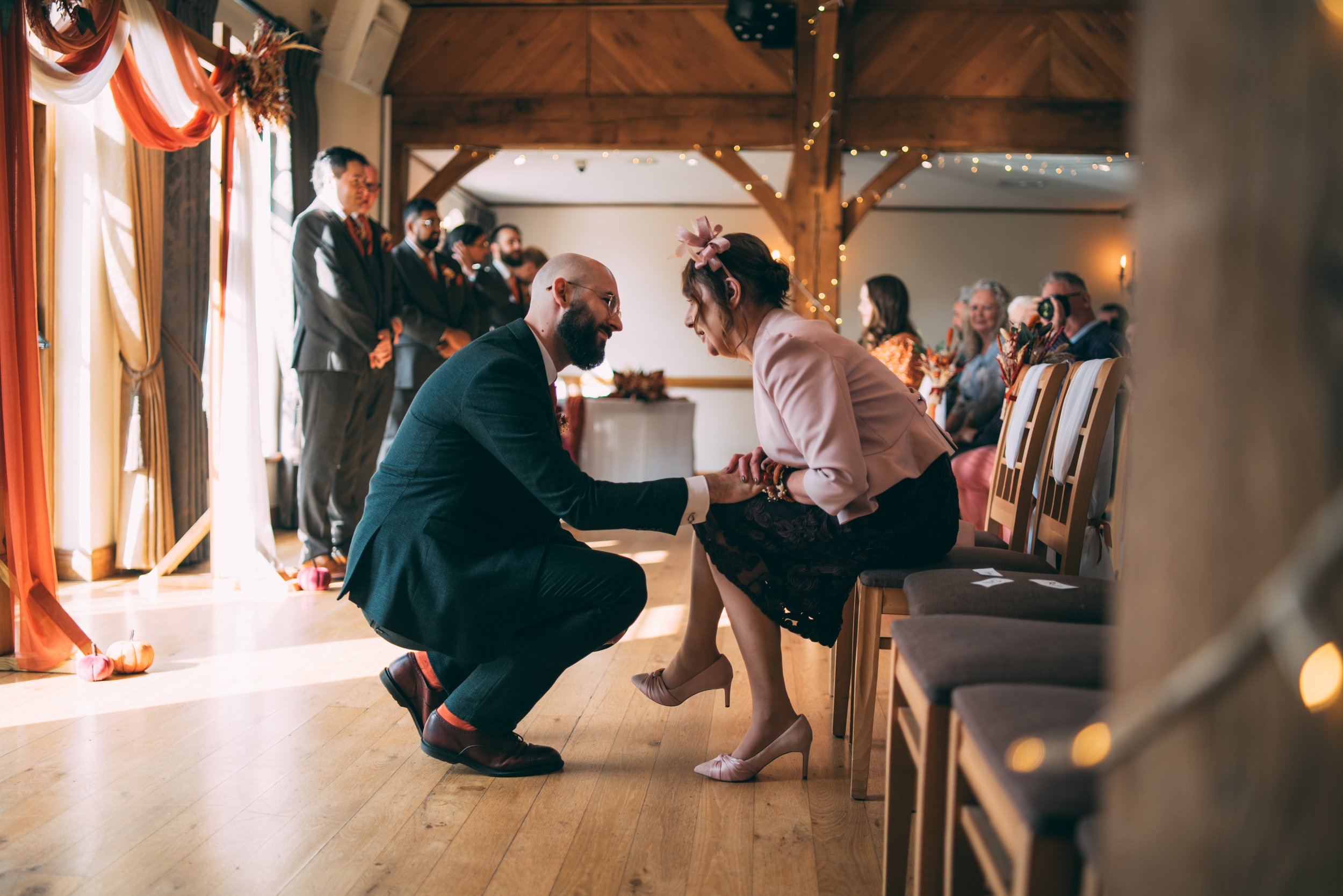 A man kneeling and holding the hands of a woman seated on a chair during a wedding ceremony inside a decorated room with string lights and guests watching.