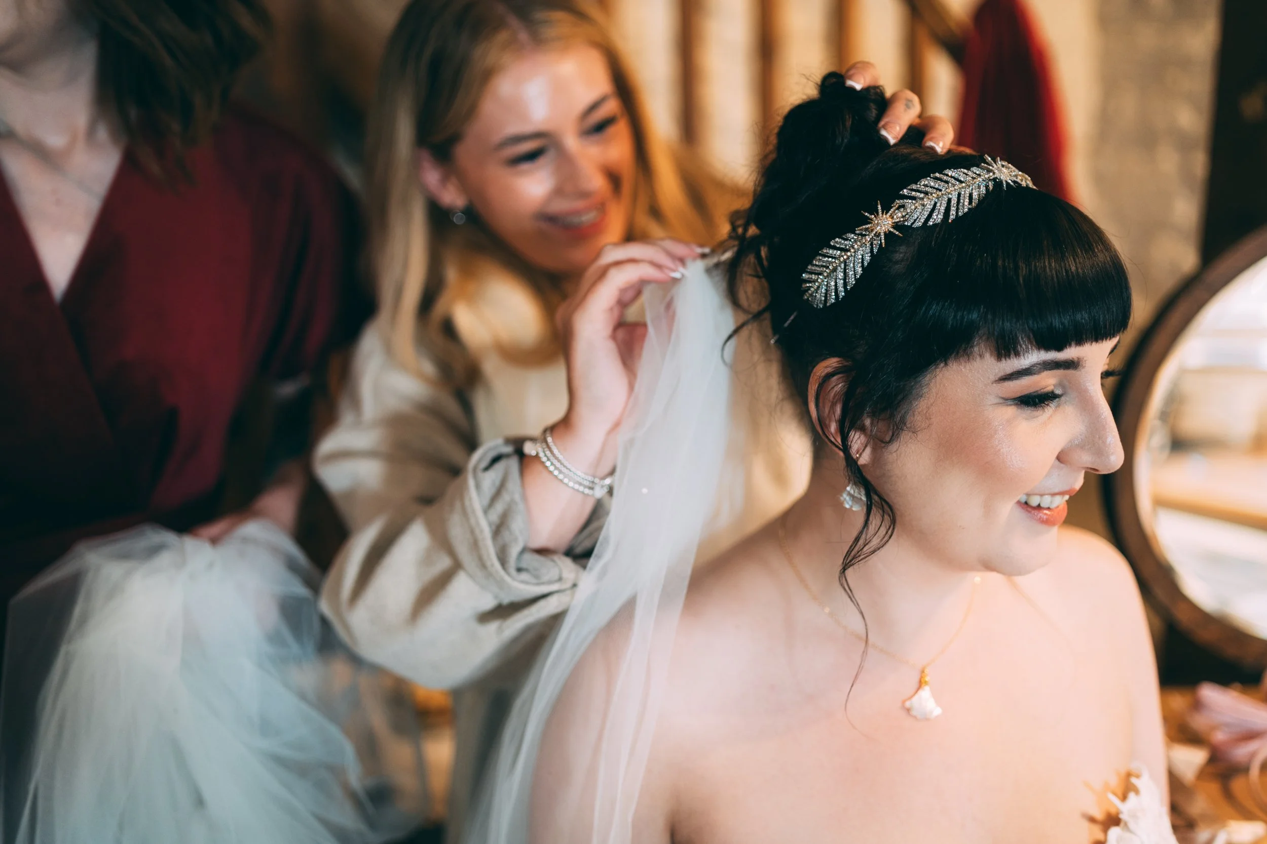 A woman with black hair and bangs, wearing a white dress and a golden necklace, smiles as she gets her veil adjusted by two women helping her, one with blonde hair and the other with brown hair, in a warmly lit room.