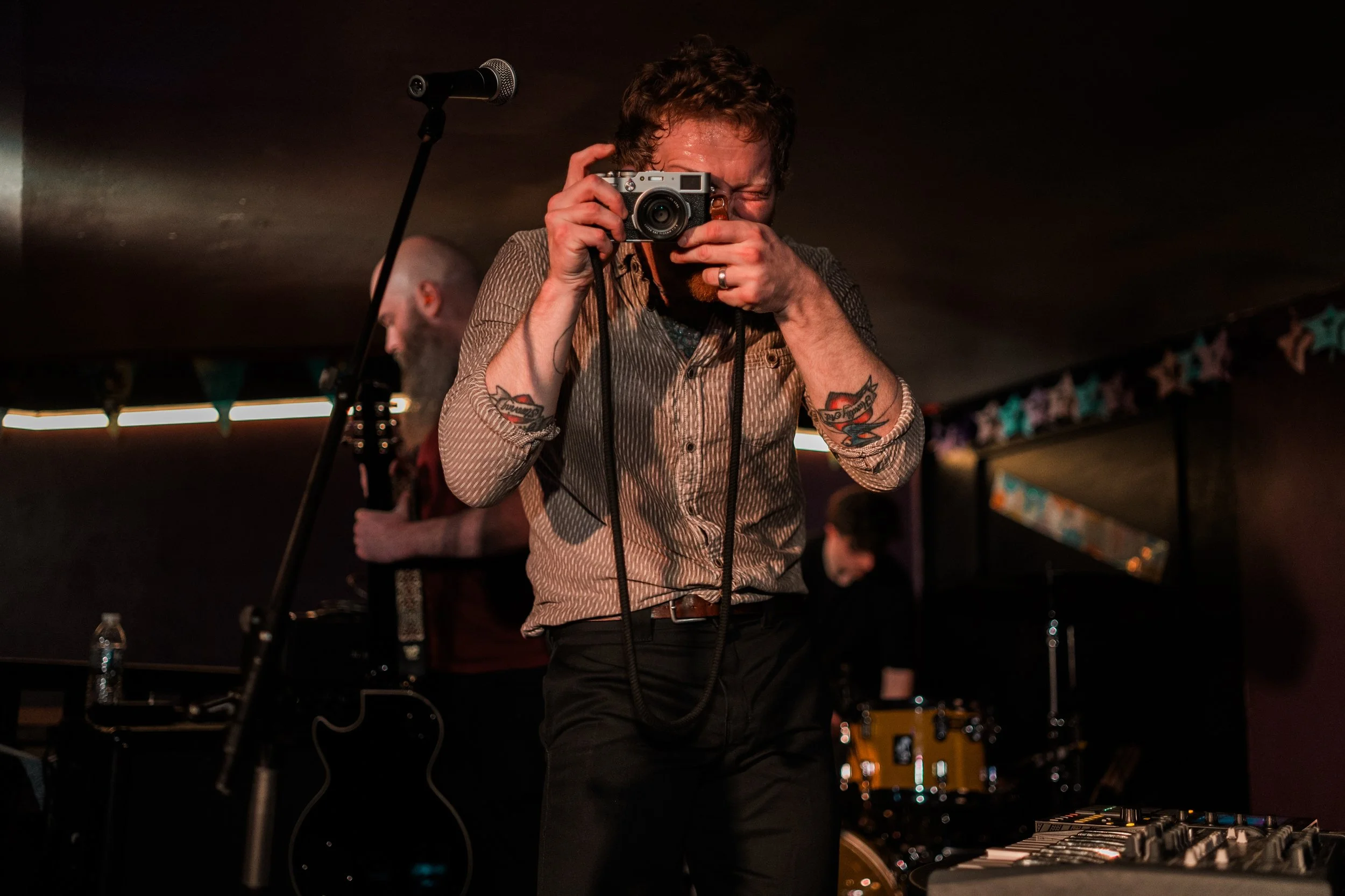 A man with curly hair and tattoos on his arms takes a photo with a camera at a music performance, with a microphone, a guitar, and other band members in the background. Promotional Music Photography. Live event photography.
