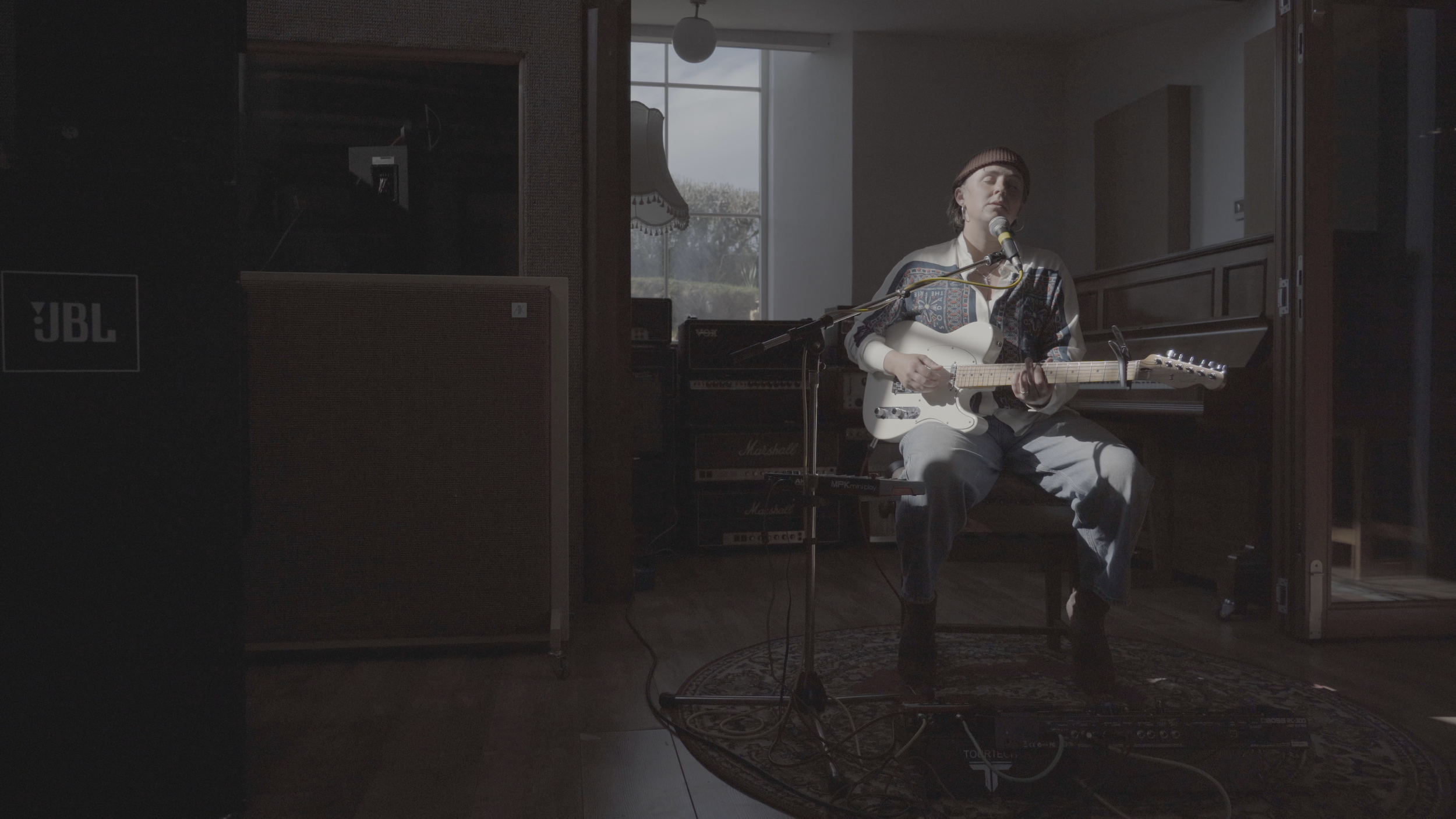 A woman sitting on a stool playing an electric guitar and singing into a microphone in a music studio, with amplifiers and a piano in the background.