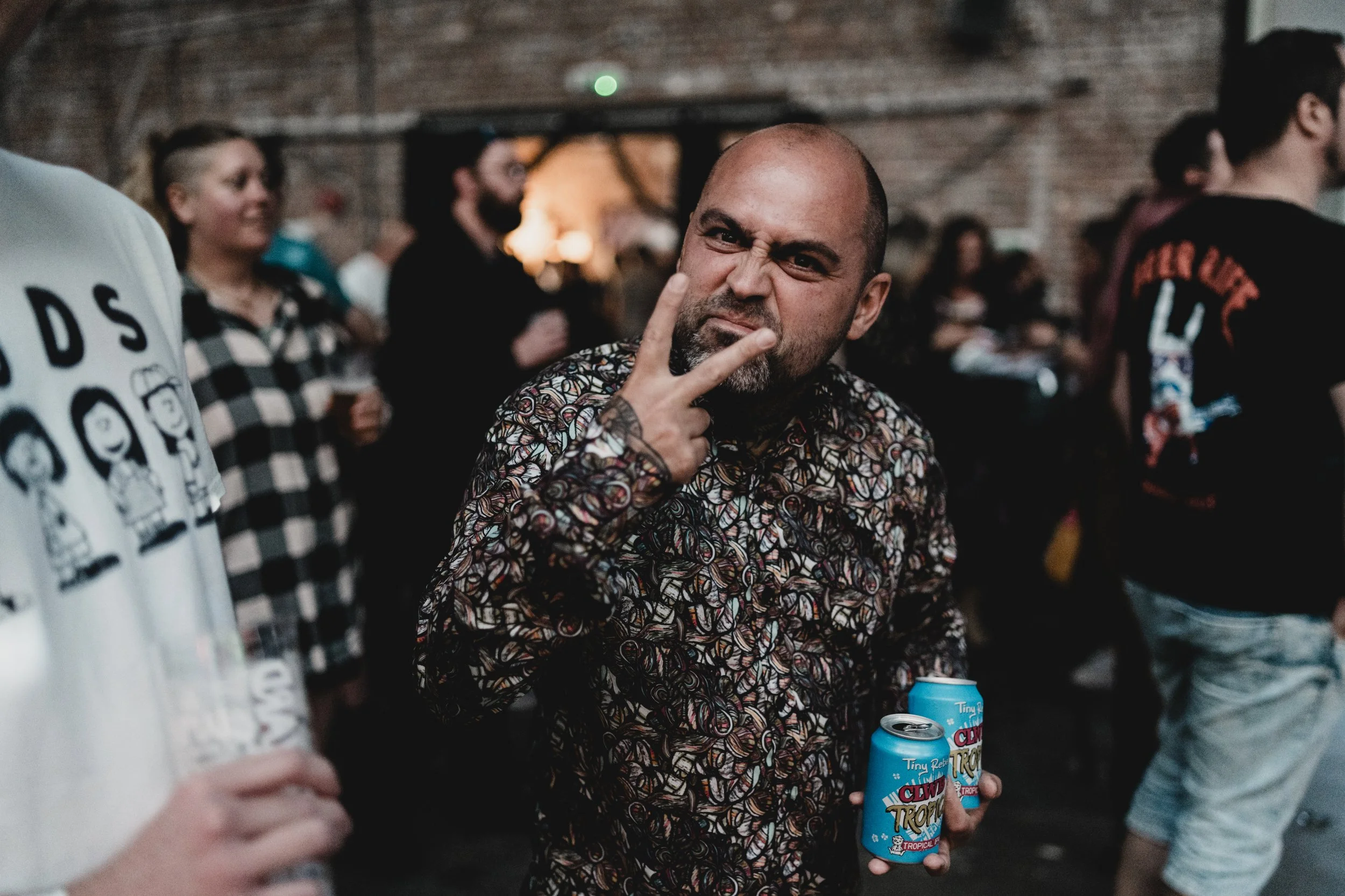 Man making a peace sign gesture, holding two cans of Clwb Tropica beverage, at a social gathering or party.