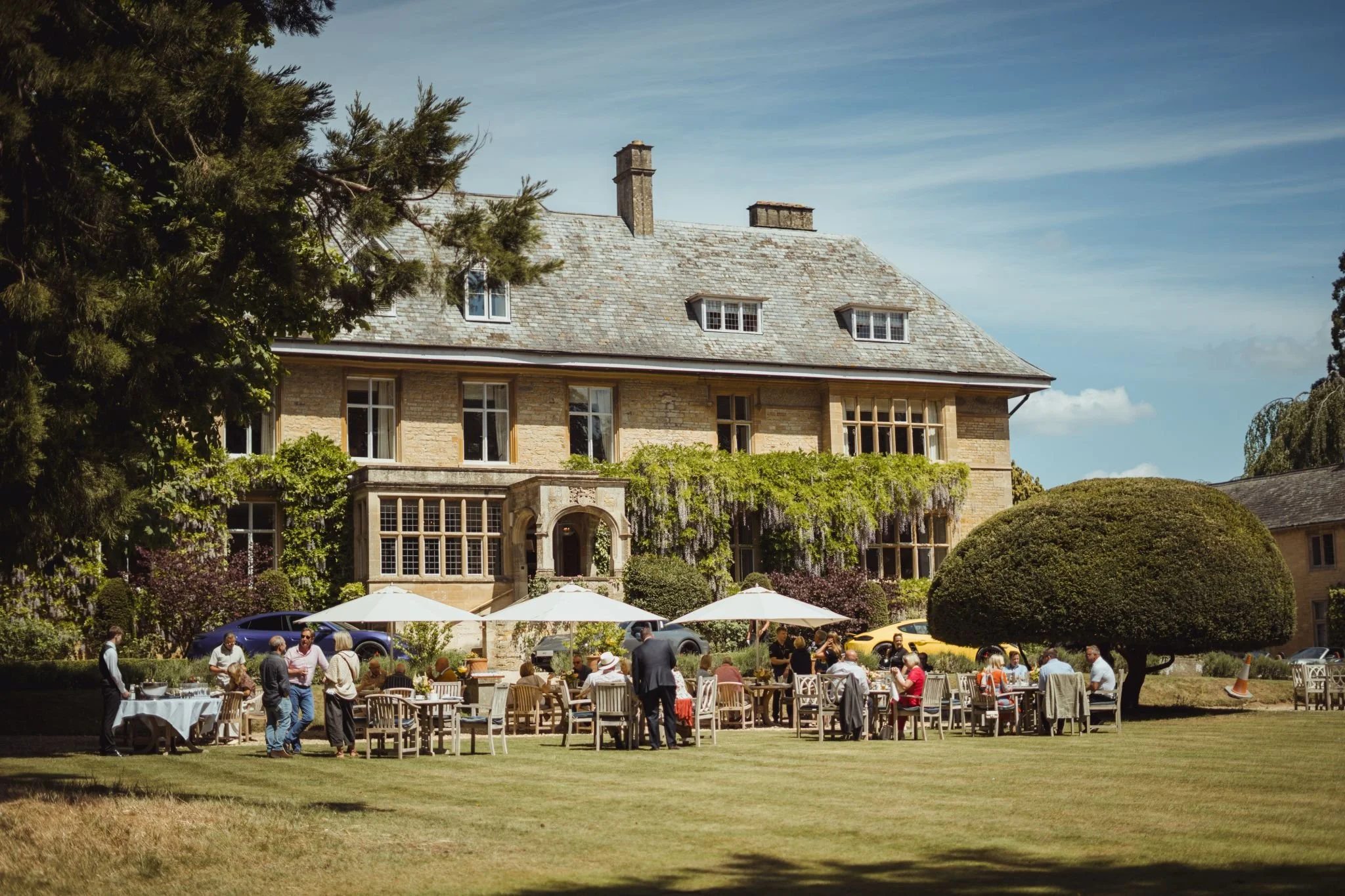 An outdoor gathering with guests seated at tables with white umbrellas in front of a large stone mansion, surrounded by trees and greenery on a sunny day.