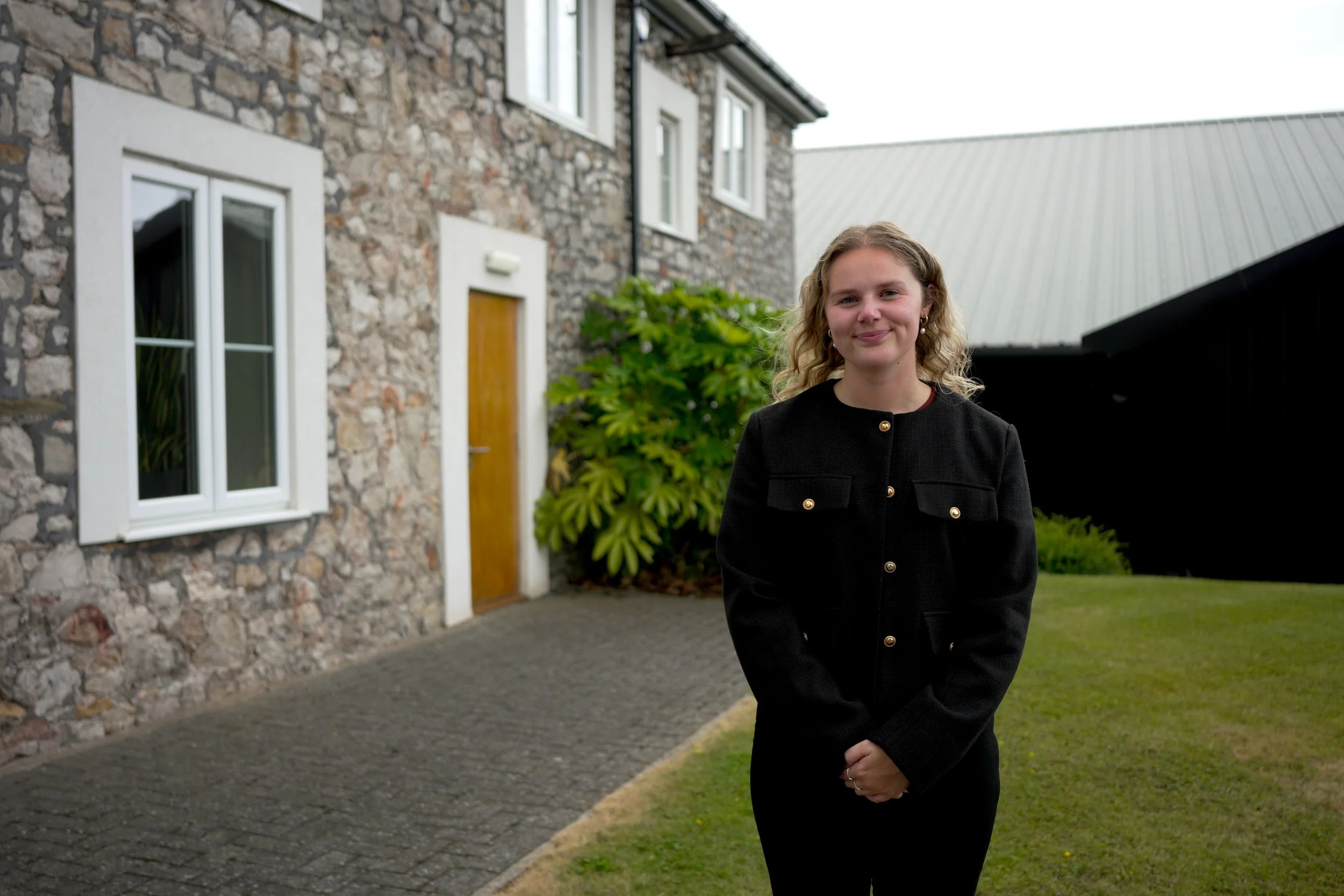 A smiling young woman with curly blonde hair wearing a black jacket with gold buttons, standing outdoors in front of a stone house with white framed windows and a wooden door, surrounded by greenery.