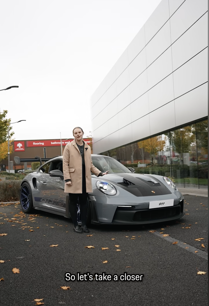 A woman standing next to a gray Porsche 911 GT3 RS parked outside a building with a reflective glass wall. The woman is wearing a beige coat and black shoes. There are fallen autumn leaves on the ground, and a red store sign is visible in the background.