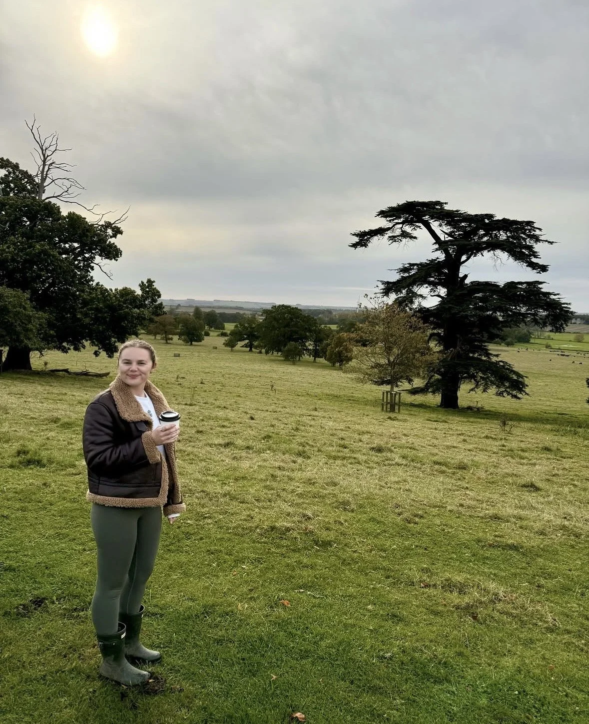 A woman standing on a grassy field holding a coffee cup, with trees and a cloudy sky in the background.