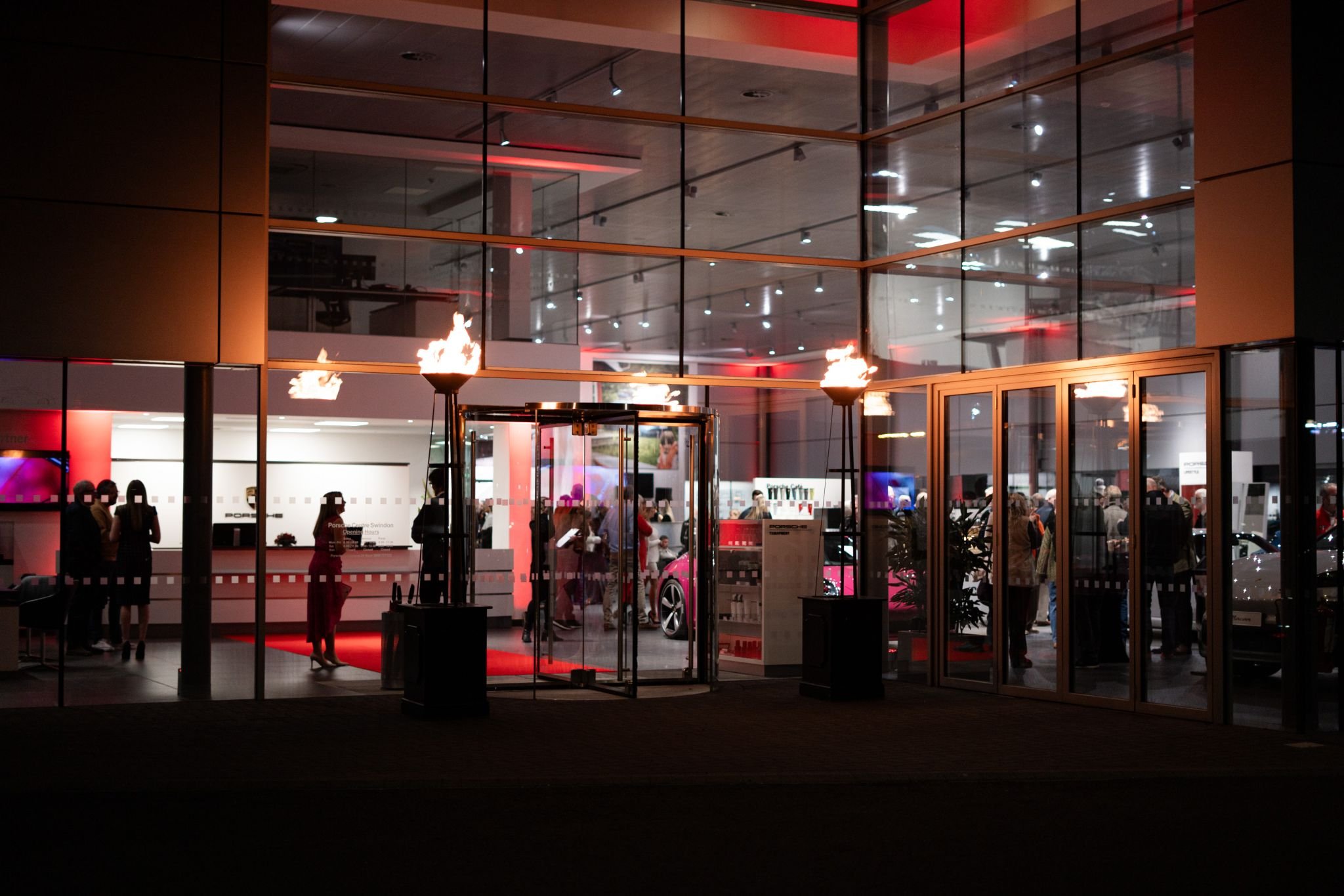 Indoor view of a car showroom lit with red lighting, featuring flames on torches on either side of the entrance, with people inside observing and walking around vehicles.