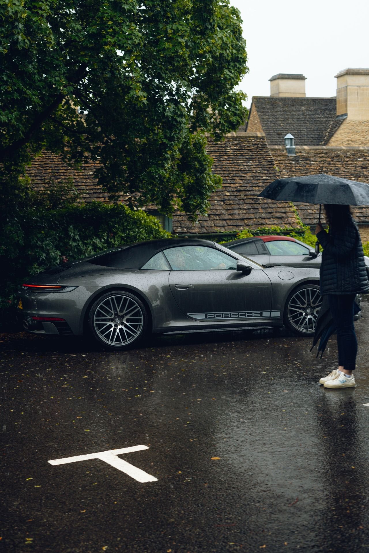 A woman holding a black umbrella standing next to a silver Porsche parked on a wet street, with rain and overcast sky.