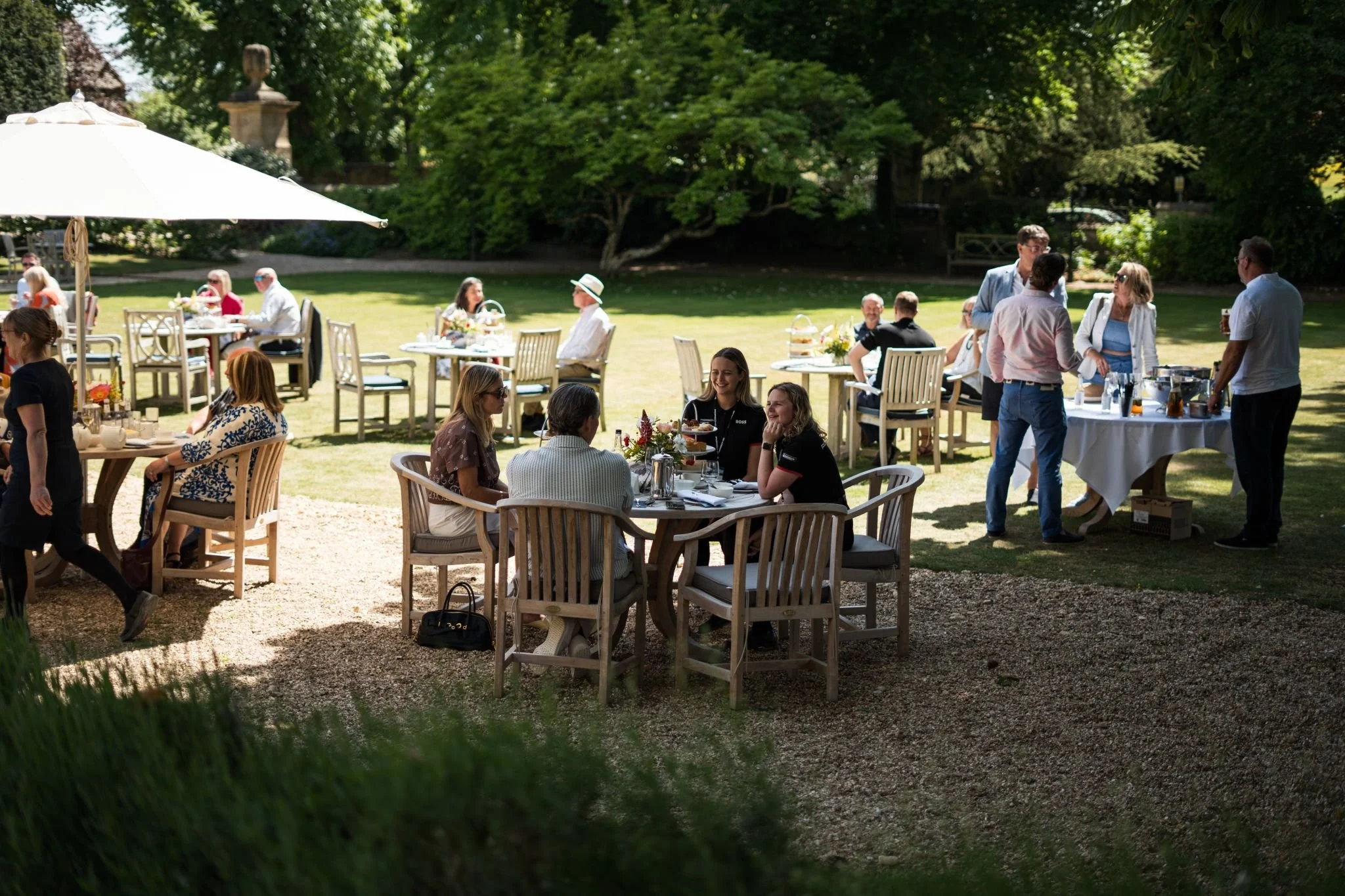 People enjoying a garden party with tables and chairs set up outdoors, some under a large umbrella, in a lush green garden with trees.