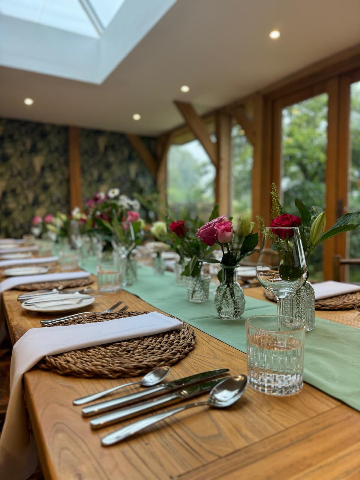 Decorated banquet table inside a wooden room with a green table runner, floral centerpieces, plates, silverware, glasses, and cloth napkins.