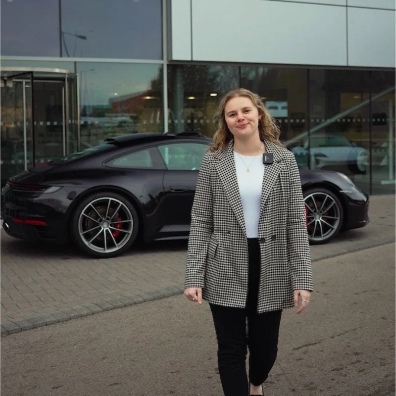 Young woman with blonde curly hair standing in front of a black Porsche outside a modern glass building.