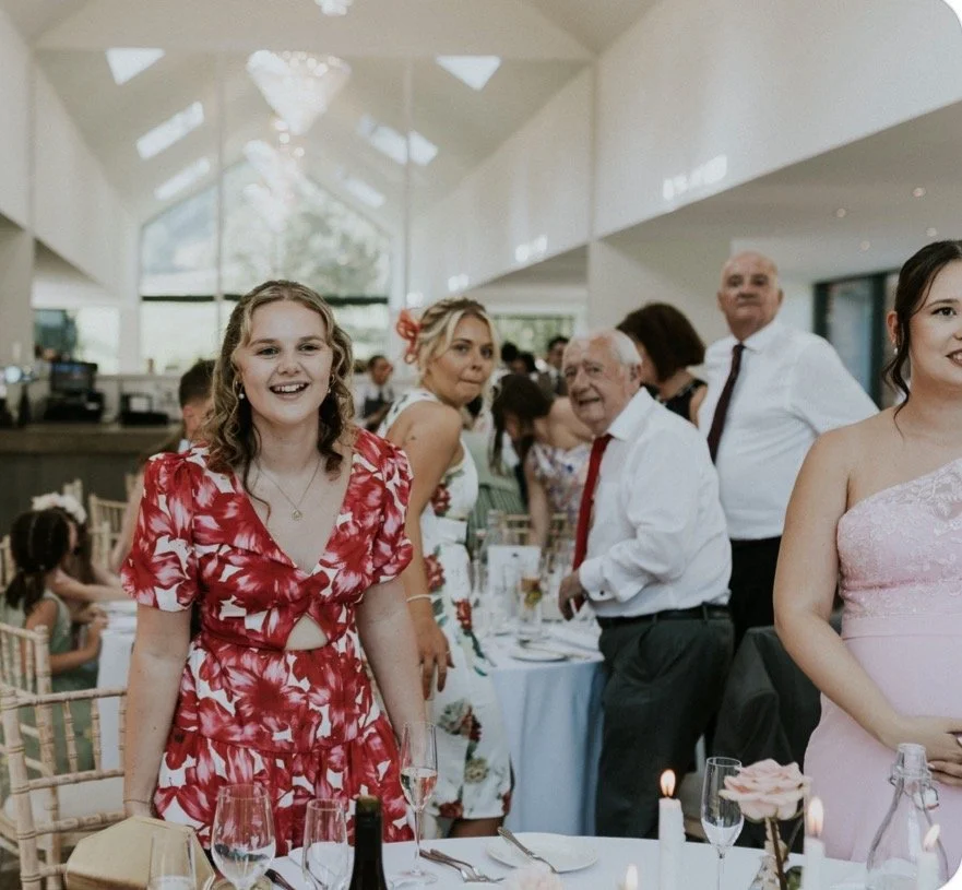 People enjoying a celebration at a bright indoor venue, with women in floral dresses and older men in white shirts, seated at a decorated table with candles and dishes.