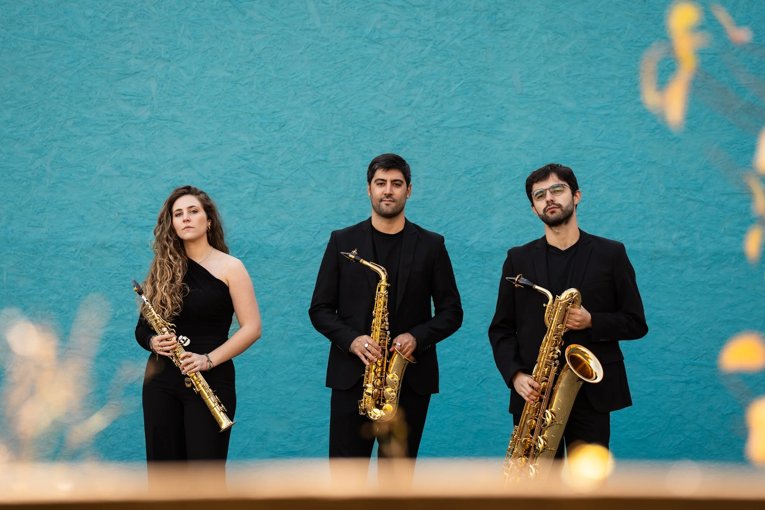 Three musicians in black suits holding saxophones, standing against a textured blue wall.
