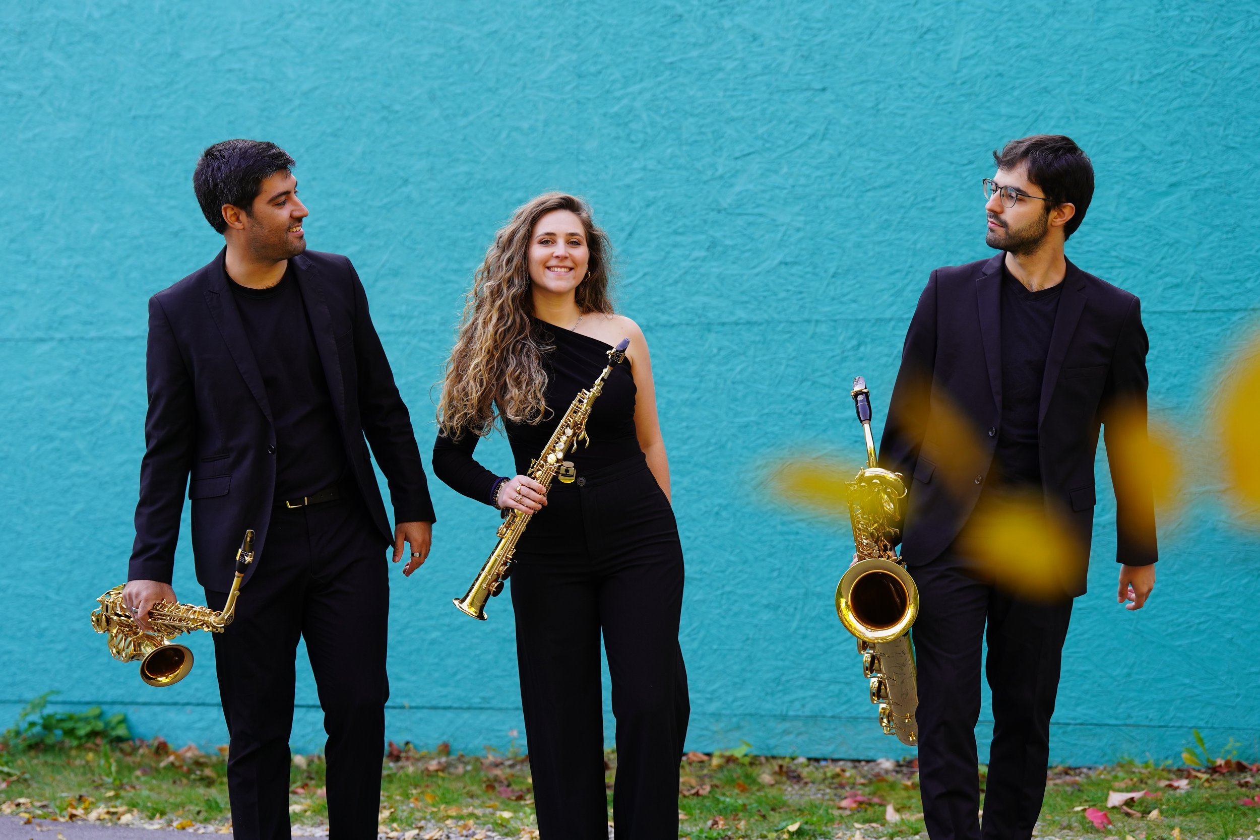 Three musicians dressed in black holding saxophones standing in front of a blue wall with leaves on the ground.