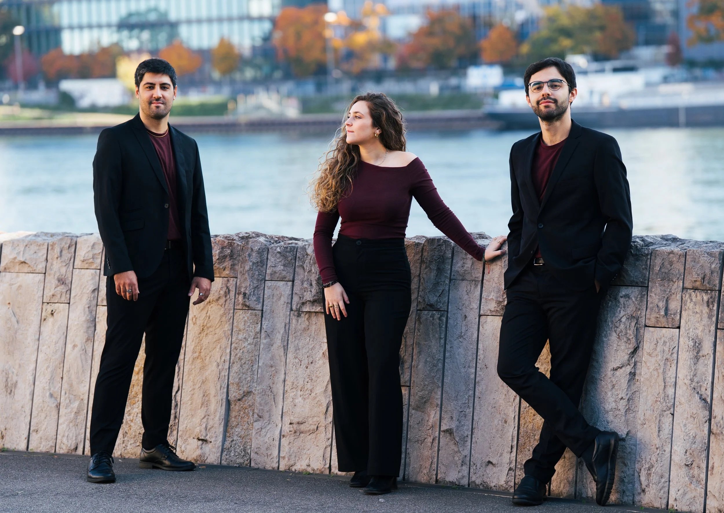Three young adults, two men and one woman, pose outdoors near a river with city buildings in the background. They are dressed in dark clothing with burgundy accents.