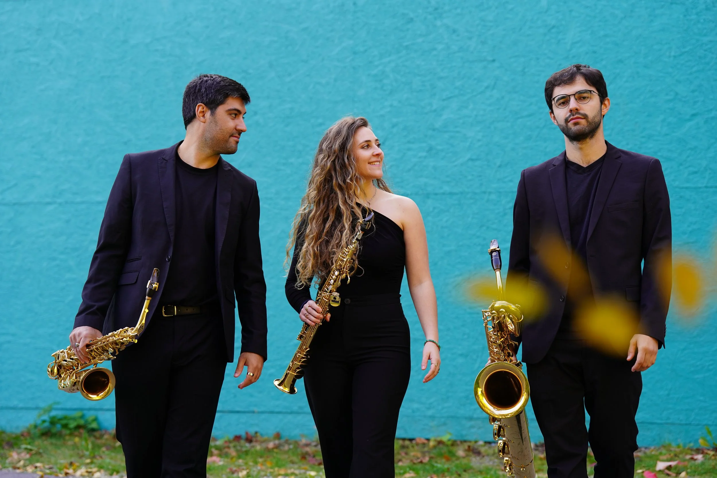 Three musicians standing outdoors against a teal wall, holding their saxophones, with the woman in the middle smiling and looking at the man on her right.