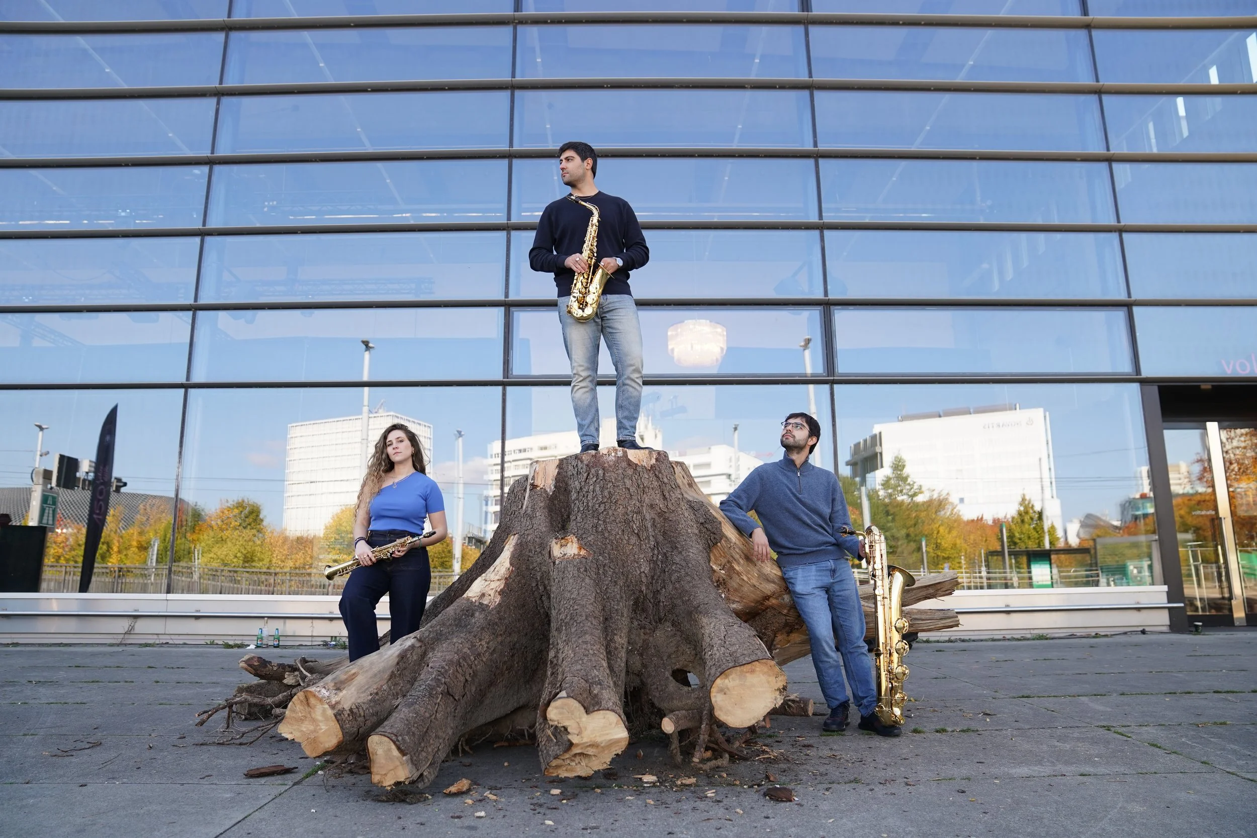 Three musicians with saxophones standing around a large tree stump in front of a glass building, with cityscape in the background.
