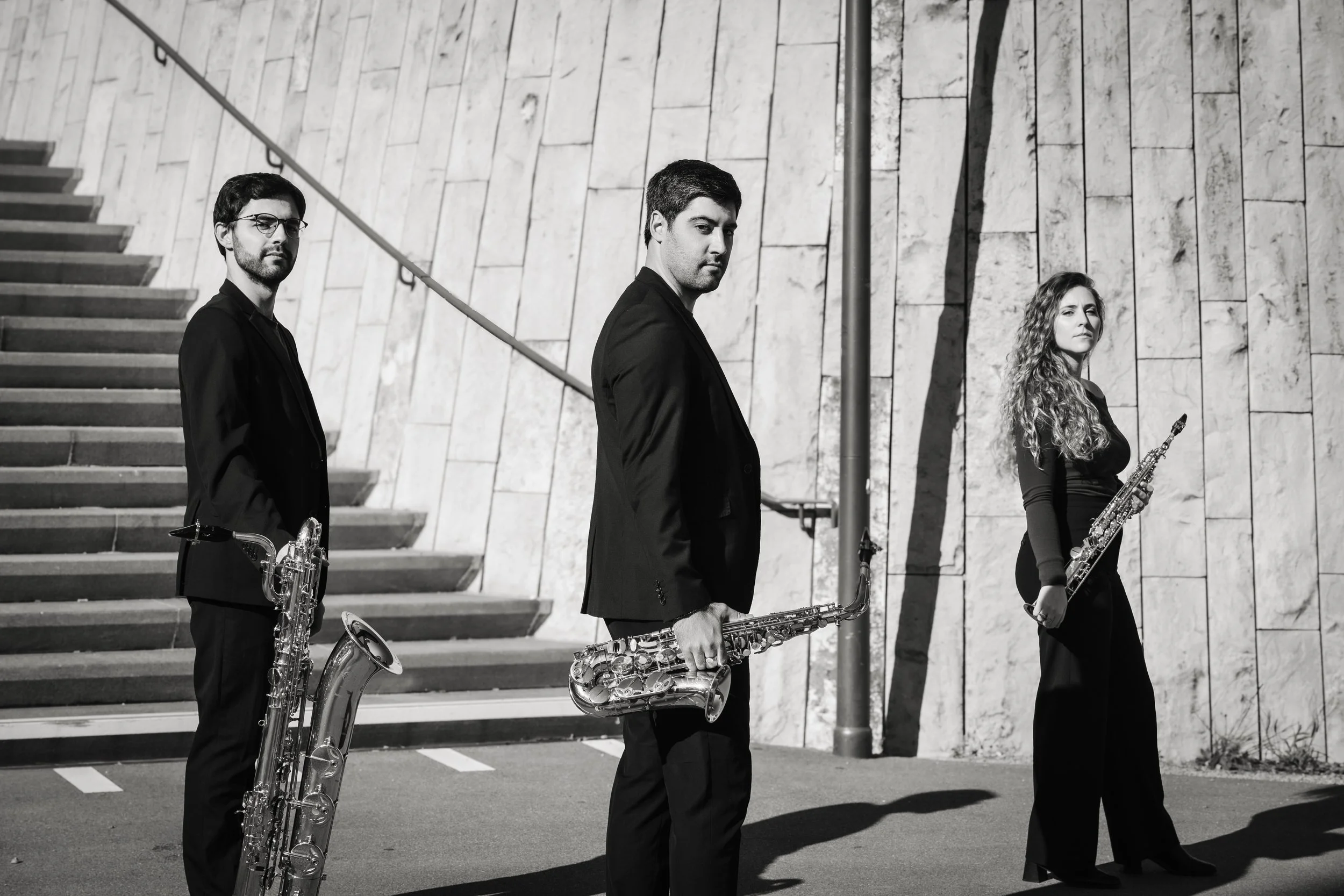 Three musicians standing outdoors in front of a stone wall, each holding a saxophone, dressed in black suits and looking at the camera.