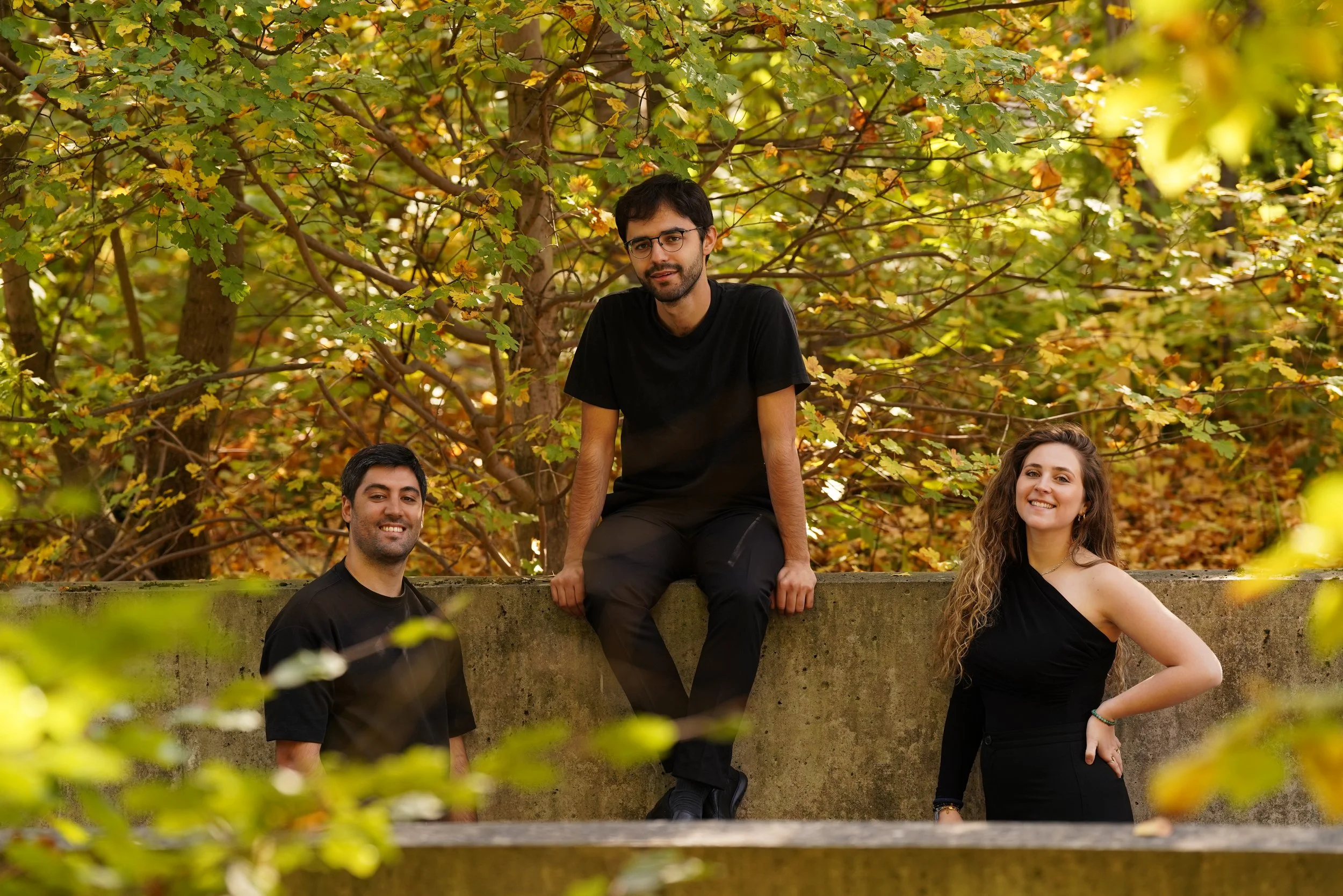 Three young adults, two men and one woman, outdoors among autumn foliage. One man is sitting on a concrete ledge, another man is standing below the ledge, and the woman is standing beside the ledge with her hand on her hip. They are smiling and dressed in casual black clothing.