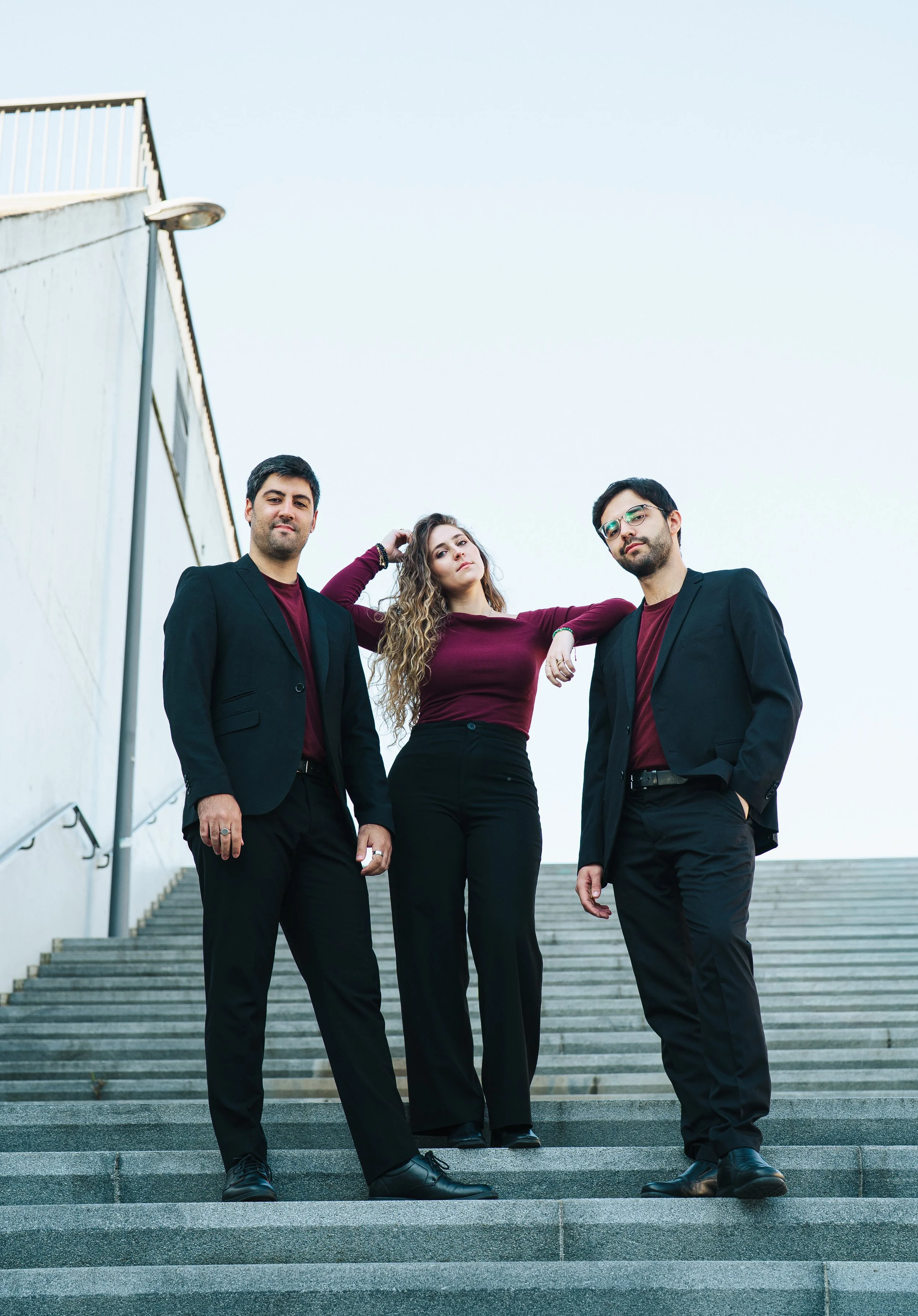 Three young adults, two men and one woman, dressed in black and burgundy clothing, standing on outdoor stairs with a clear sky background.